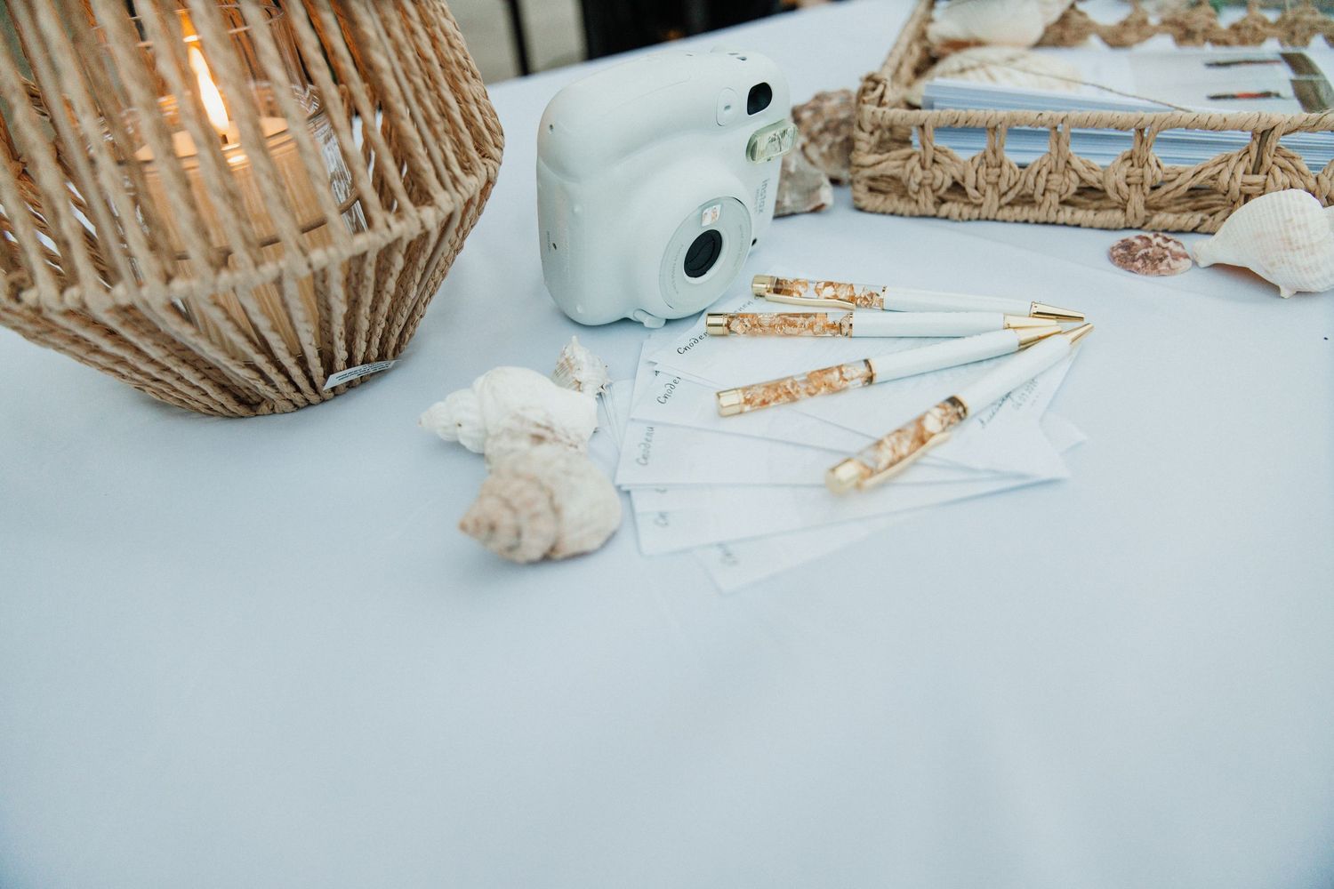 Beach wedding table with Instax camera, seashells, gold pens, and wicker decor on a light blue tablecloth.
