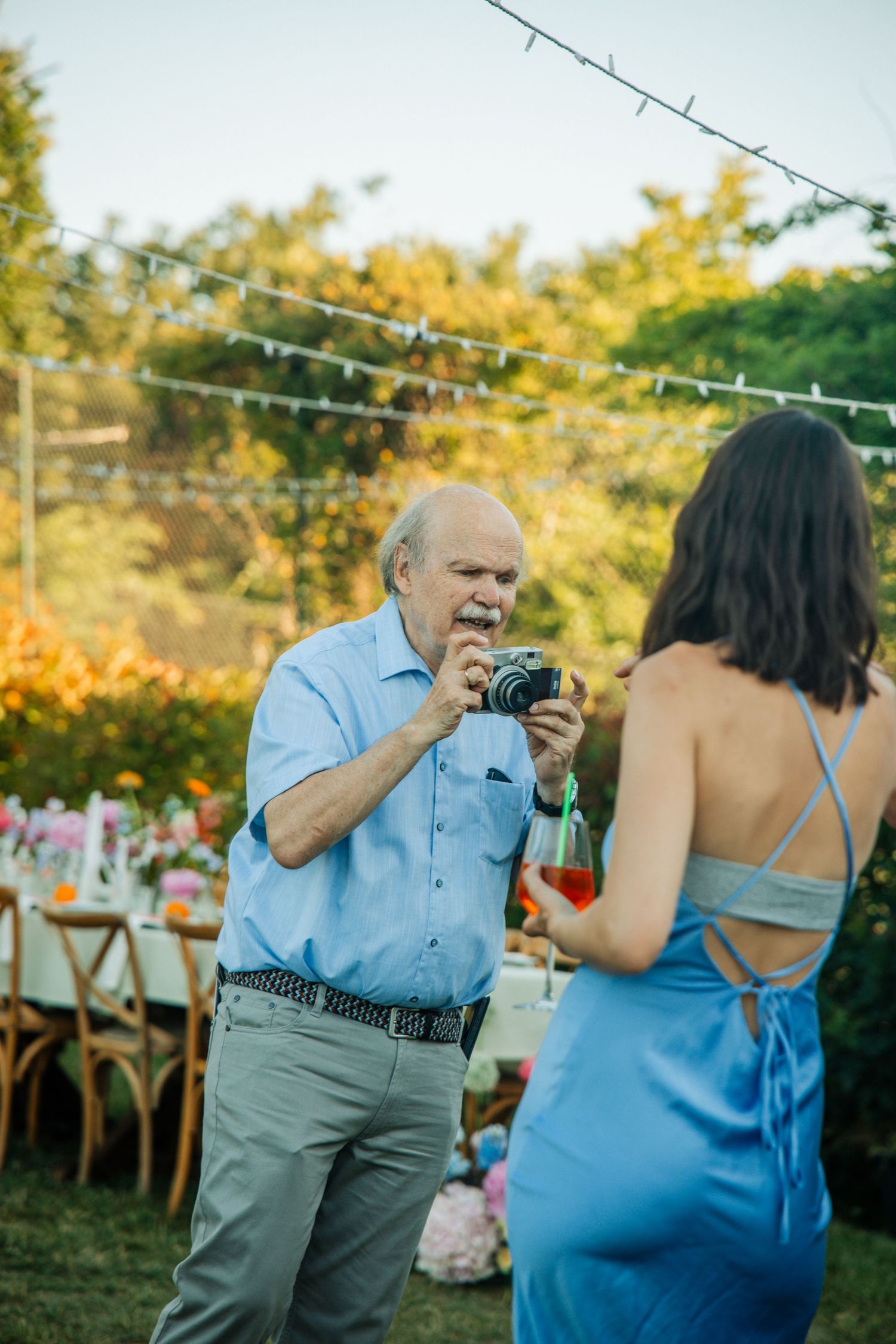 An elderly man photographs a woman in a blue dress at an outdoor garden wedding reception with string lights.