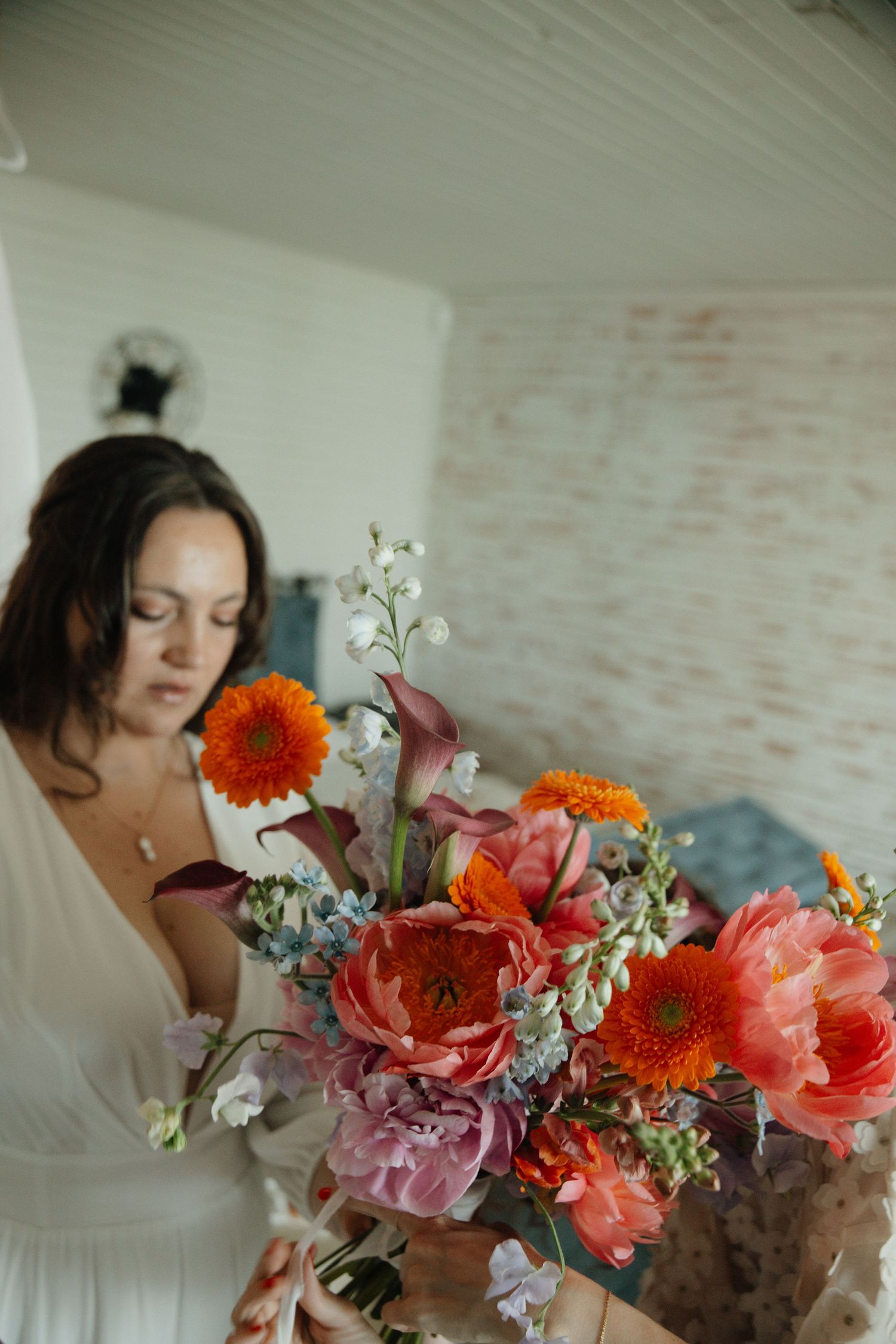 Bride in white dress holds vibrant bouquet of orange, pink, and blue flowers indoors.