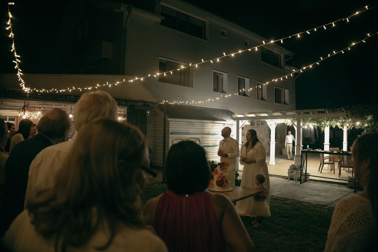 Wedding guests watch a ceremony under glowing string lights outside a white house at night.