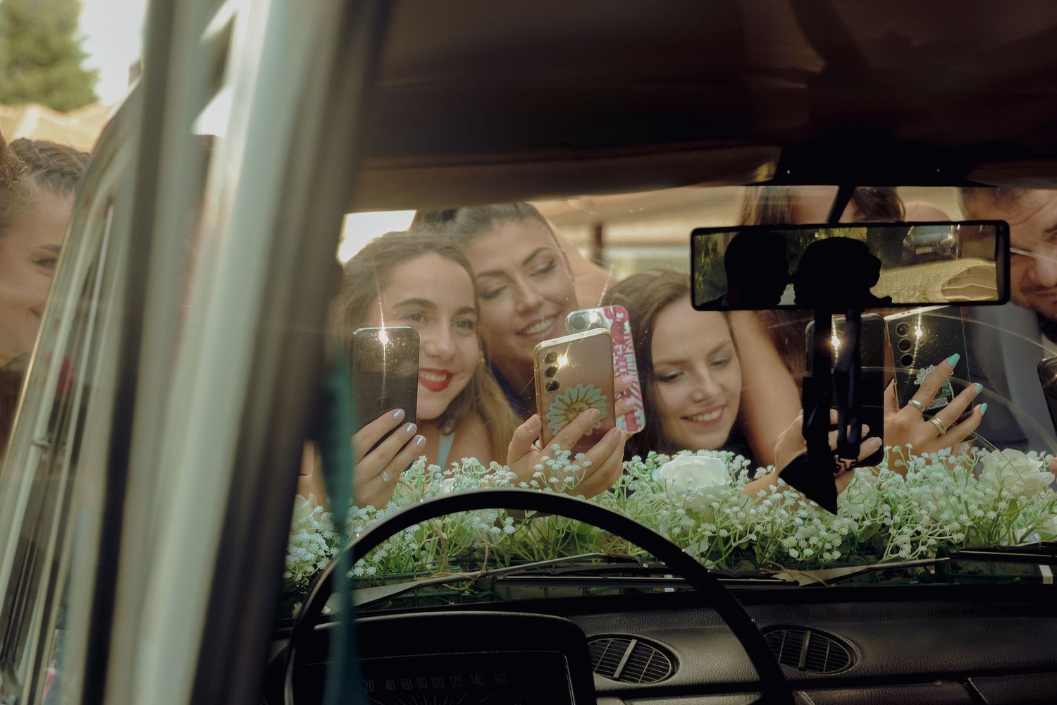 Group of young women taking selfies with phones through a car window, adorned with white flowers.