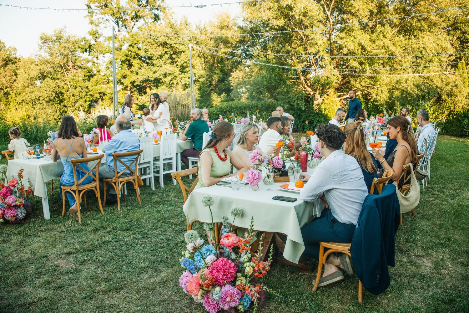 Guests seated at decorated outdoor wedding reception tables surrounded by colorful floral arrangements on green lawn.