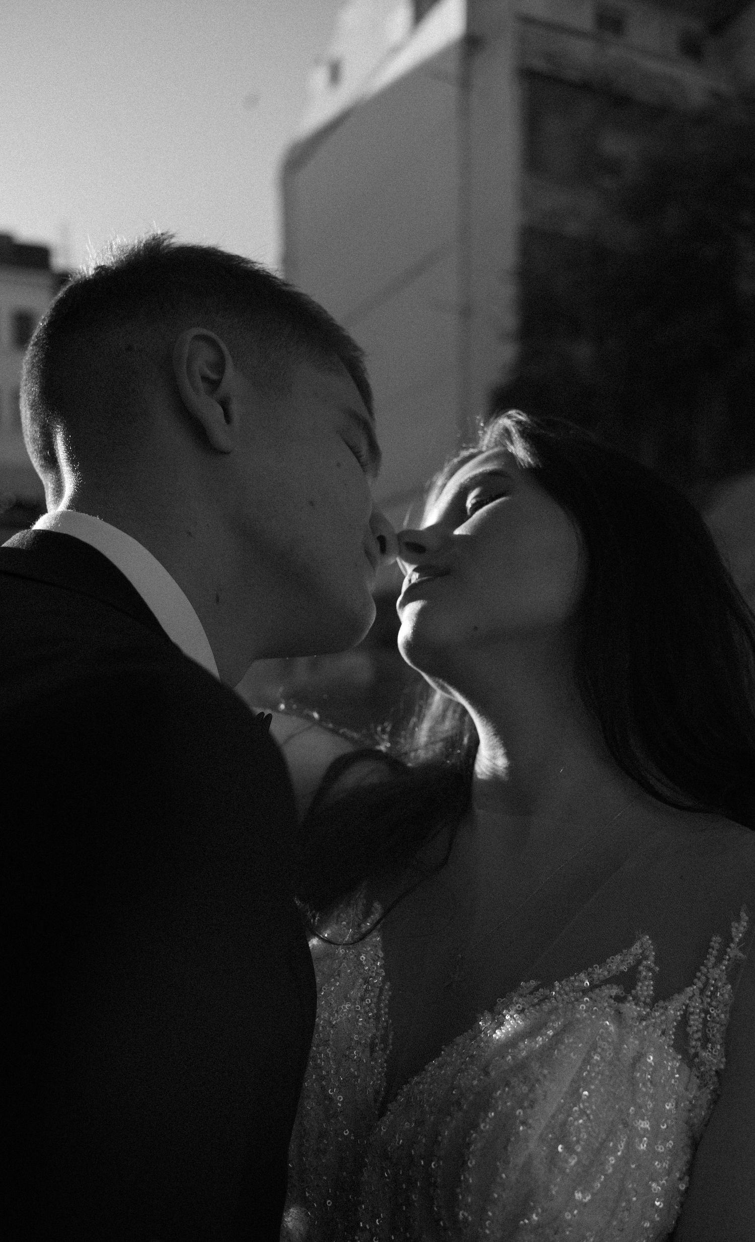 Black and white photo of a couple sharing a romantic kiss, bride in beaded gown and groom in suit outdoors.
