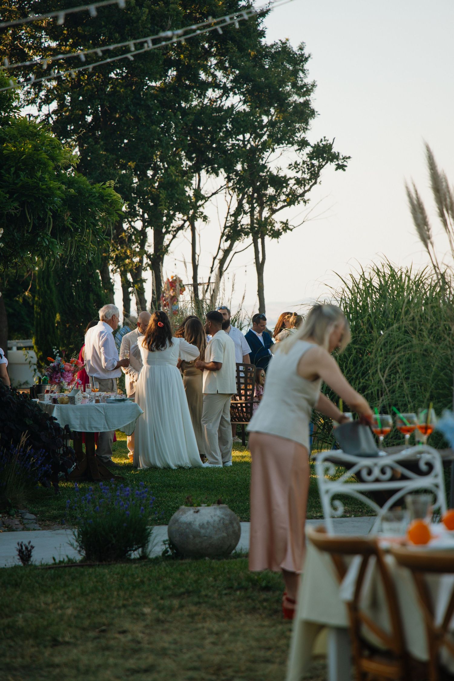 Wedding guests mingling outdoors at dusk near elegantly set tables with string lights and tall grass.