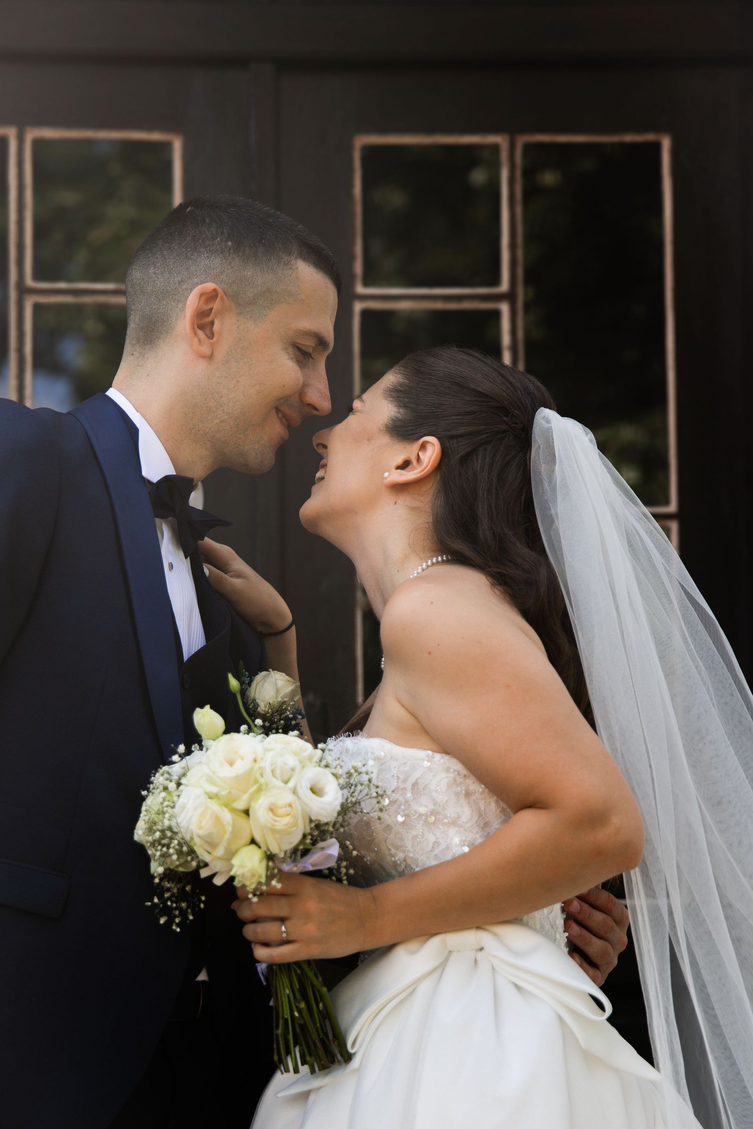 Bride and groom sharing a romantic moment, nearly kissing, bride holding white rose bouquet in elegant wedding attire.