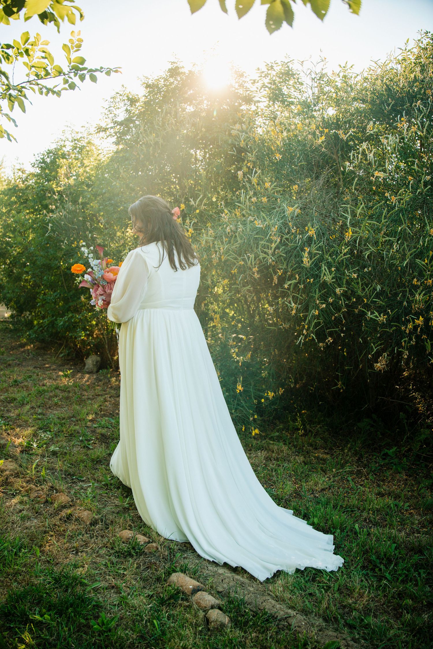 Bride in white flowing gown holding bouquet, standing in sunlit garden with lush greenery.