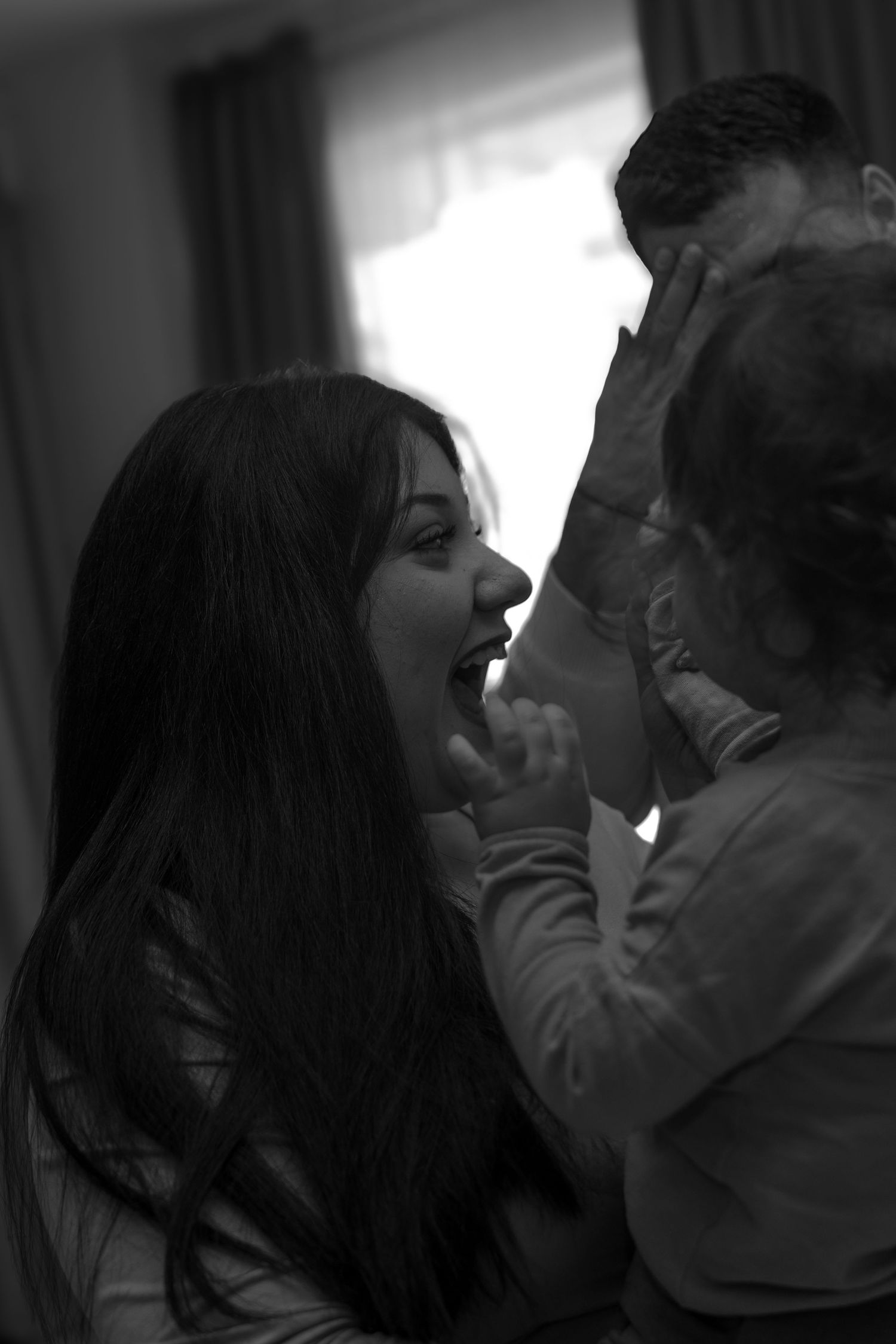 Black and white photo of a laughing woman with long dark hair playing with a toddler, man visible in background.