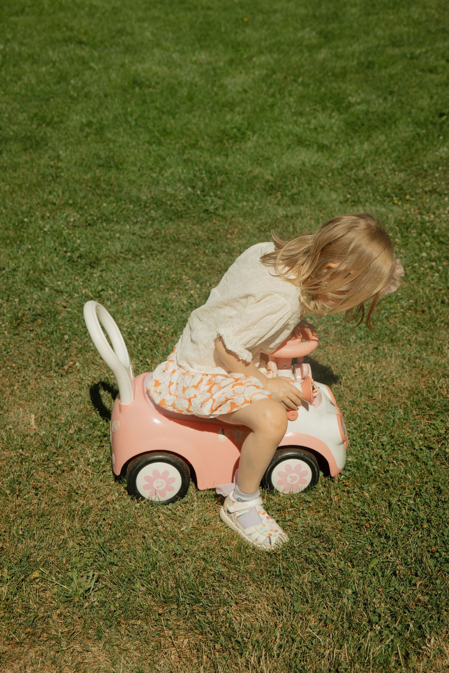 Toddler girl with blonde hair riding a pink toy car on green grass outdoors.
