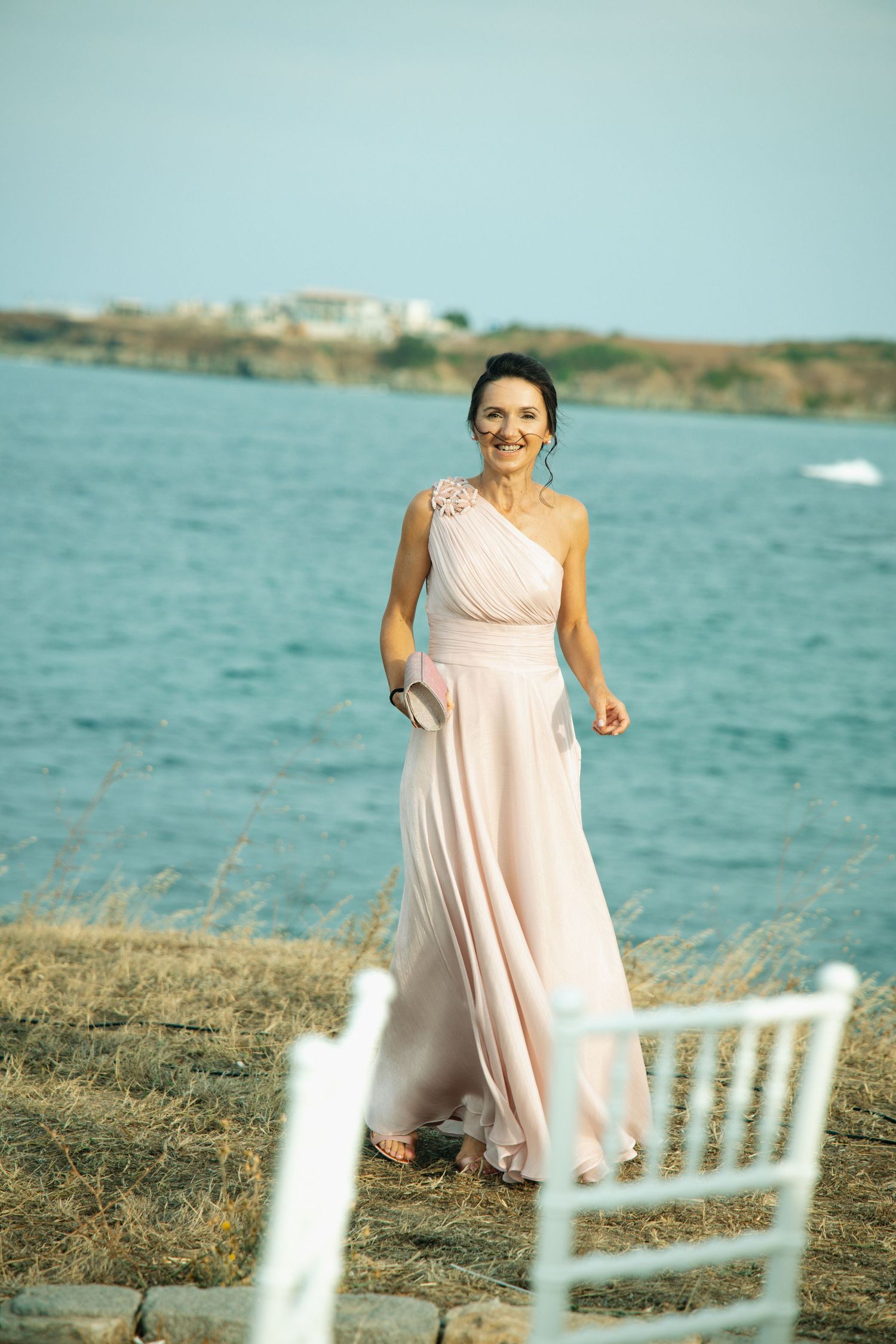 Woman in elegant pink one-shoulder gown posing by seaside at outdoor wedding event.