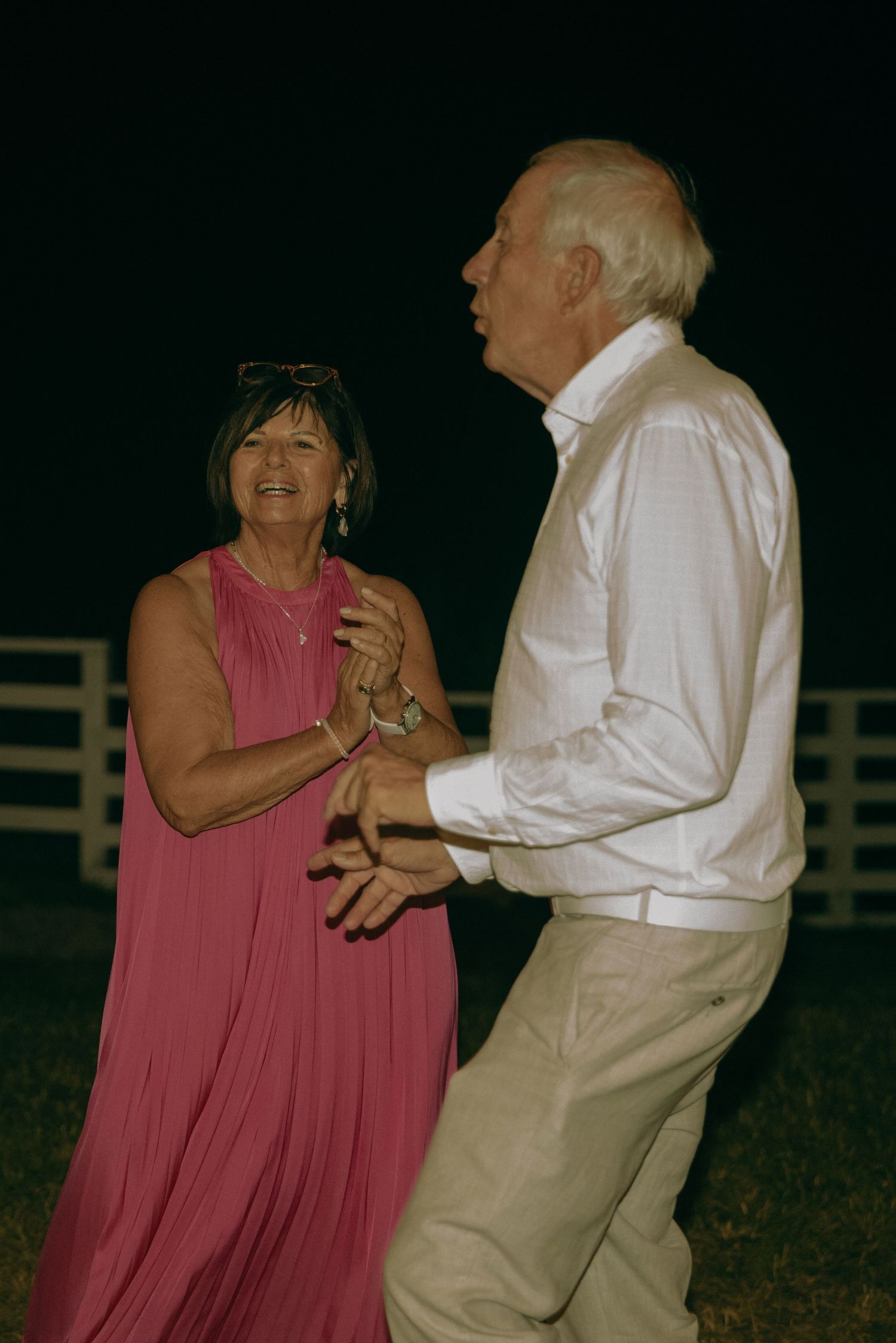 Elderly couple dancing outdoors at night, woman in pink dress, man in white shirt and khaki pants.