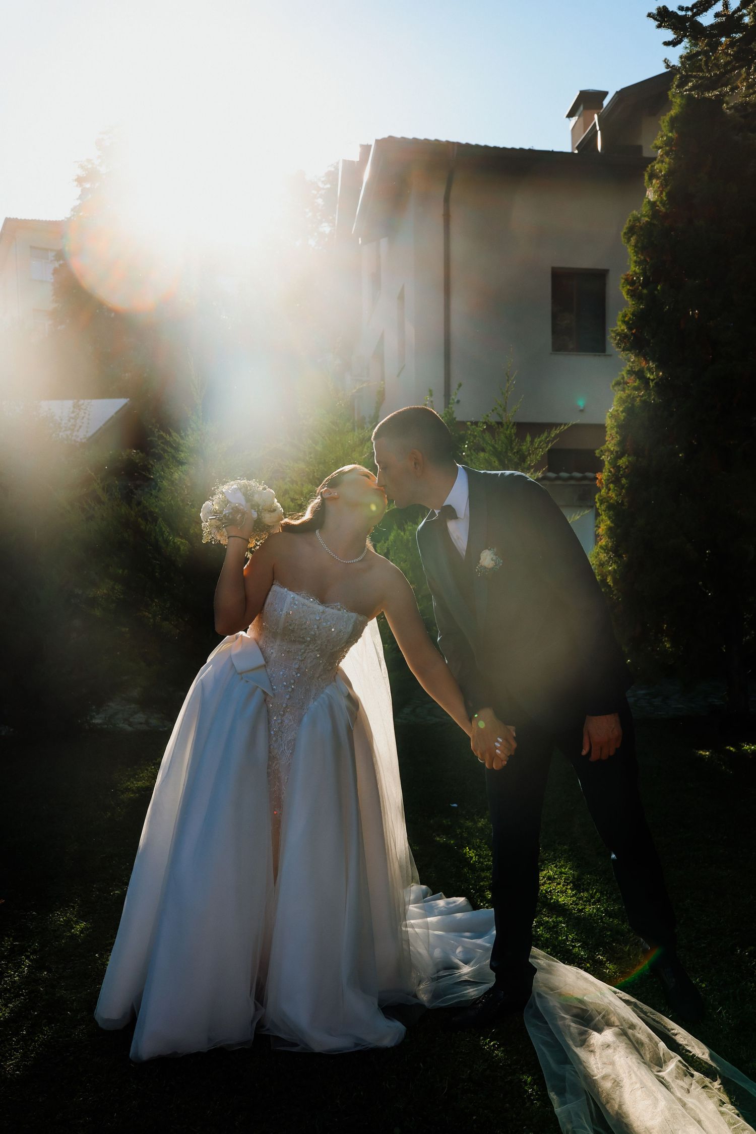 Bride and groom kissing while holding hands in sunlit garden, bride wearing strapless ball gown holding bouquet.