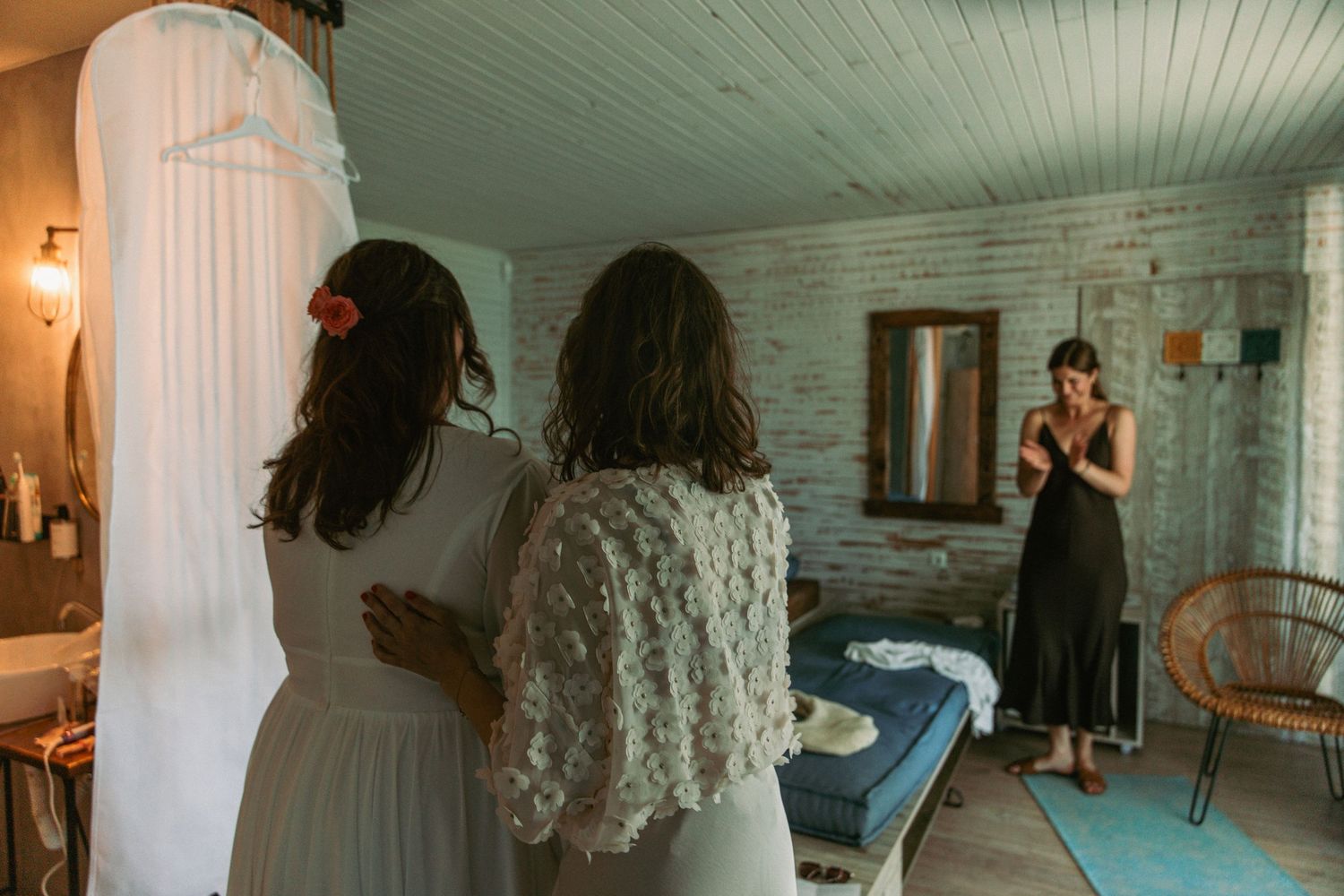 Bride and bridesmaids prepare in a rustic room as a woman in black dress looks on admiringly.