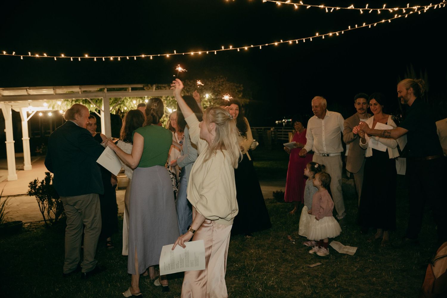 Wedding guests dancing outdoors at night under warm string lights with sparklers at evening reception.