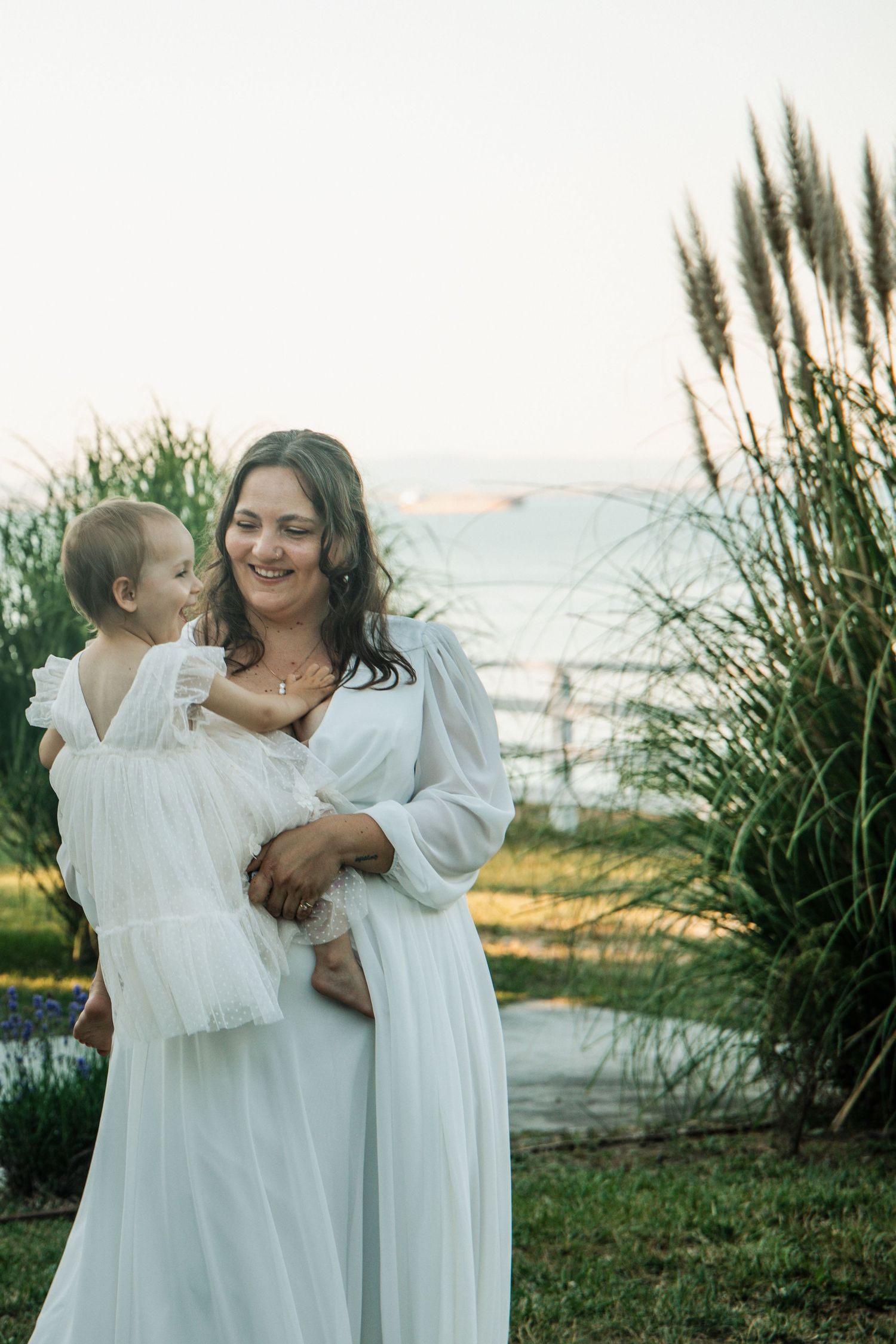 A smiling woman in a white dress holds a baby in a white outfit outdoors near tall grass and water.