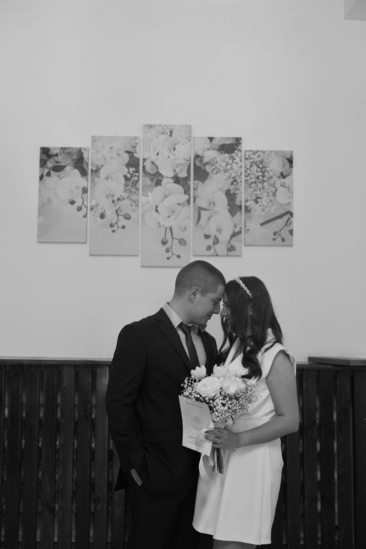 Black and white photo of newlyweds sharing intimate moment, bride holding bouquet, floral art on wall.
