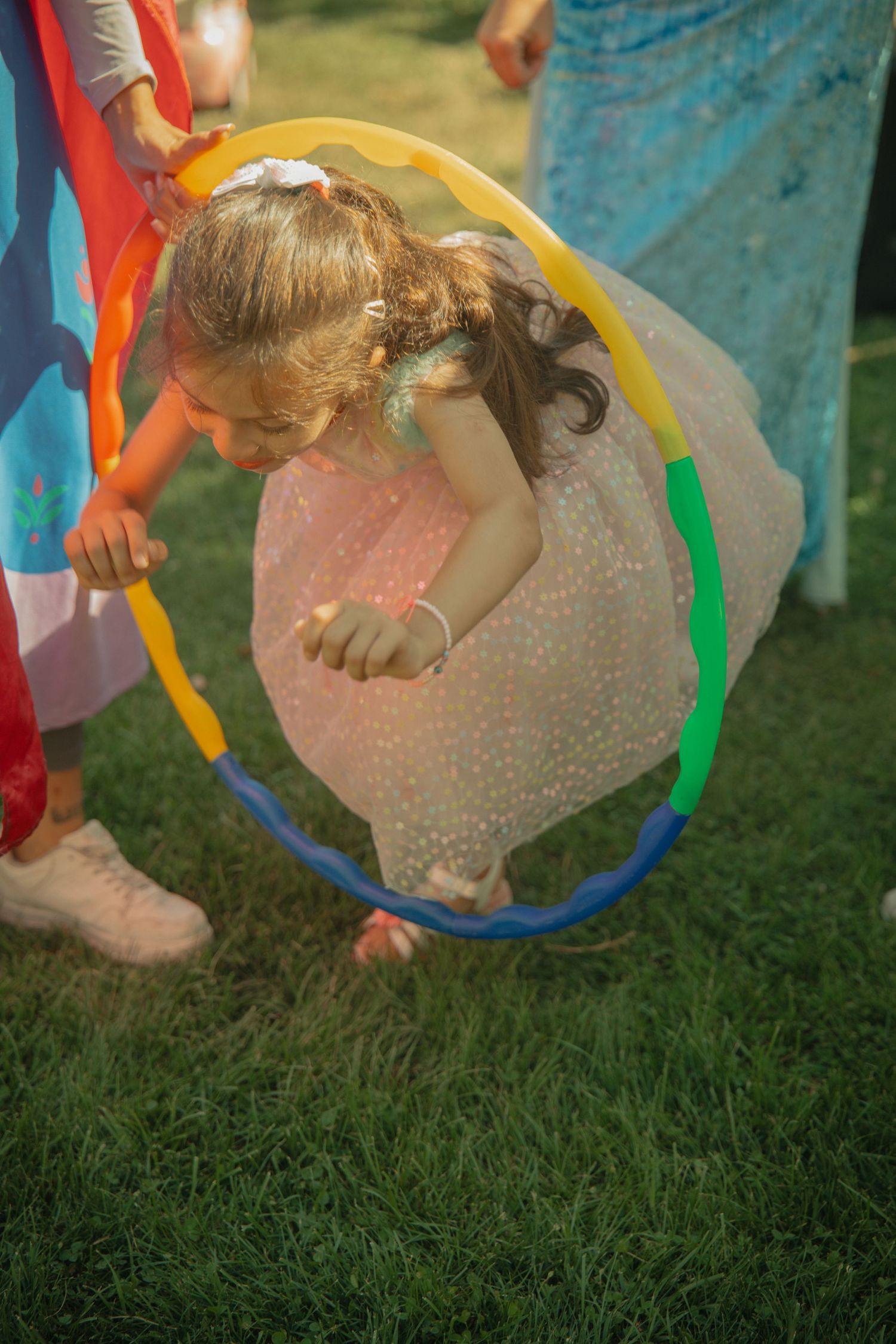 Young girl in pink dress crawling through colorful hula hoop on green grass at outdoor children's party.