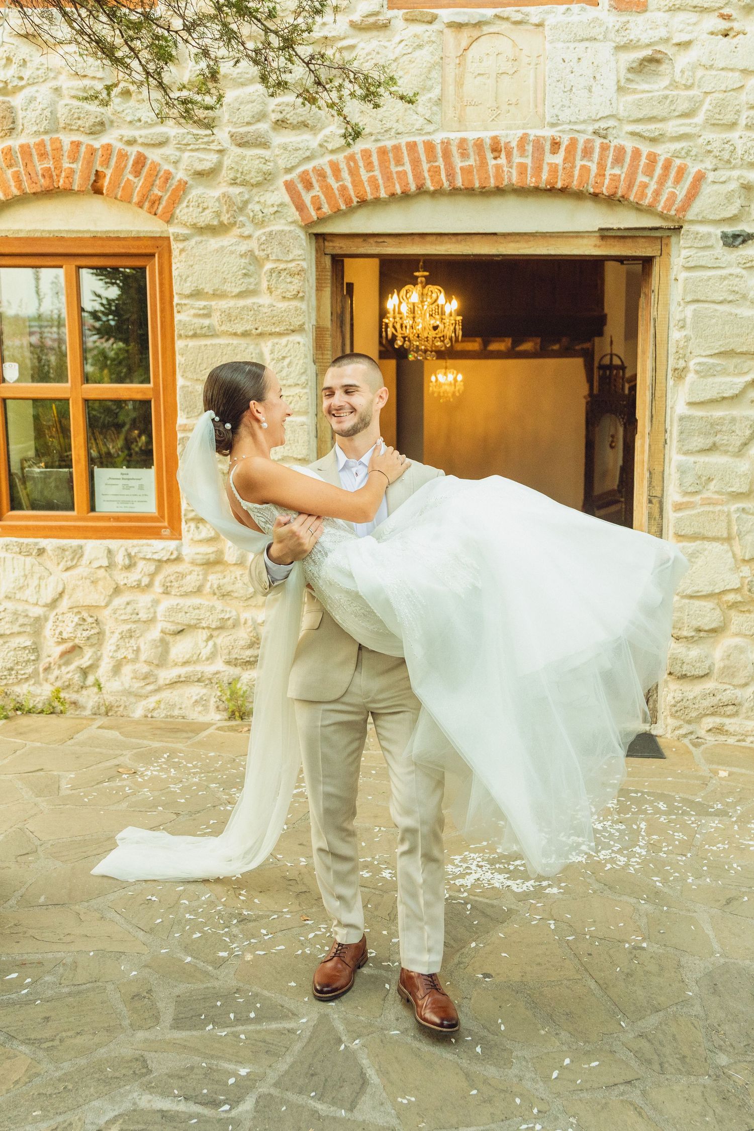 Groom in beige suit carries bride in white wedding dress outside rustic stone building with chandelier.