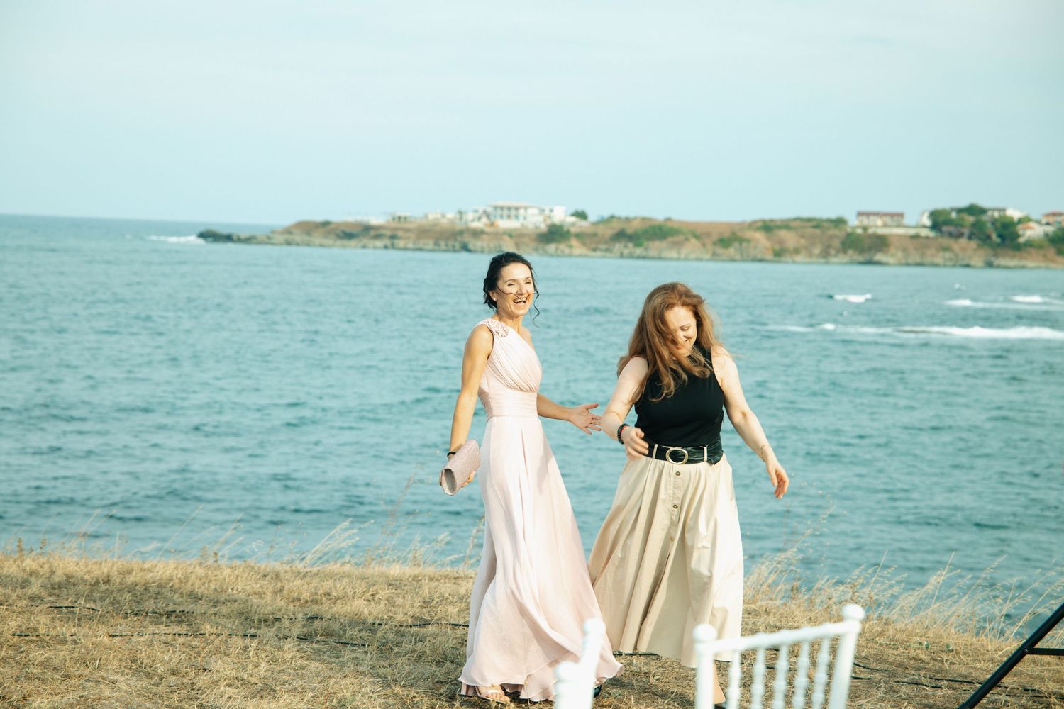 Two women in elegant dresses holding hands and walking along a scenic coastal cliff on a sunny day.