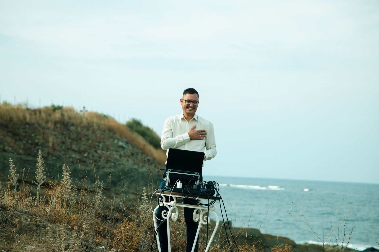 Man in white shirt standing outdoors by the sea with a laptop on a tripod stand on a rocky hillside.