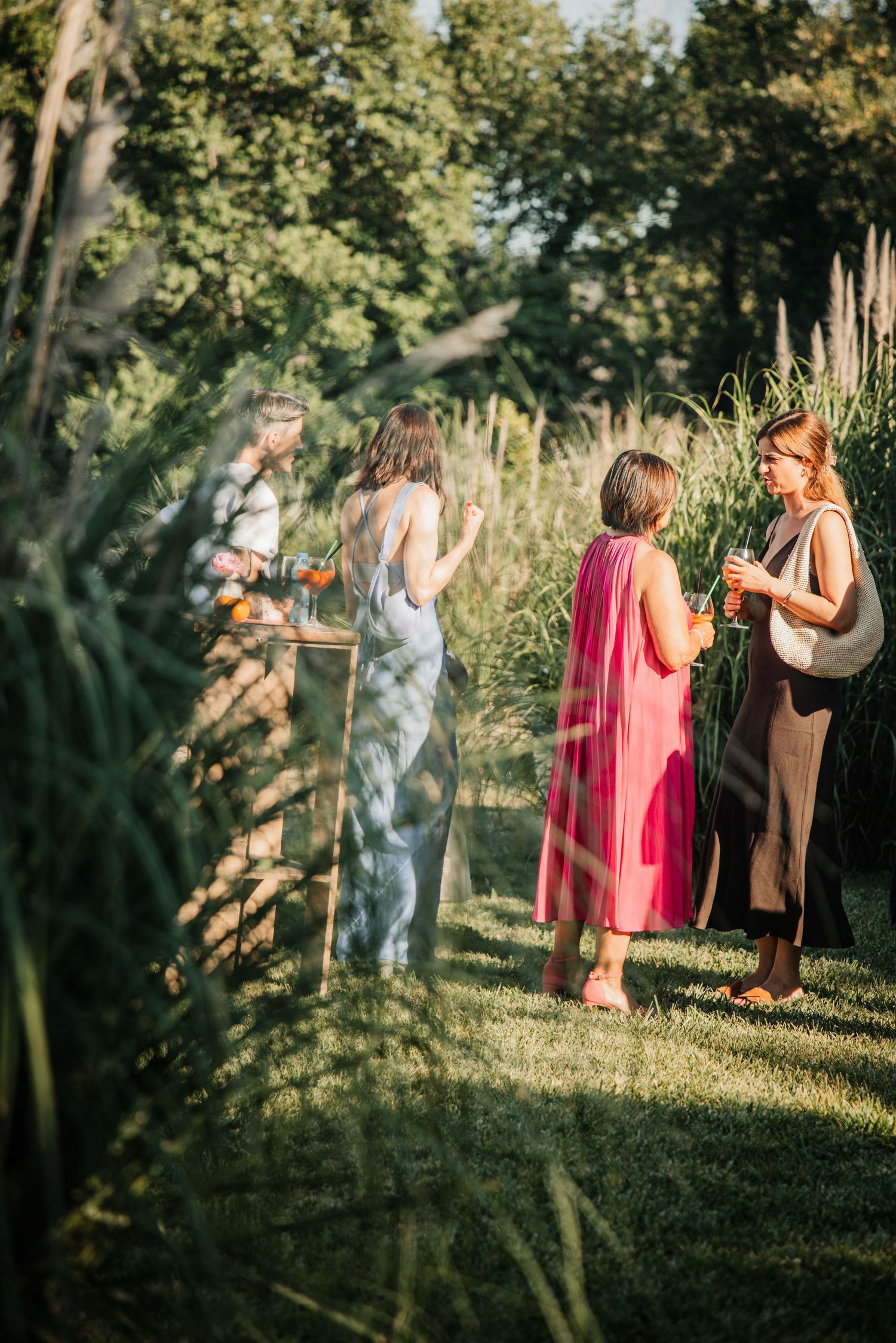 Four elegantly dressed women in colorful gowns socialize outdoors at a garden party in warm evening light.