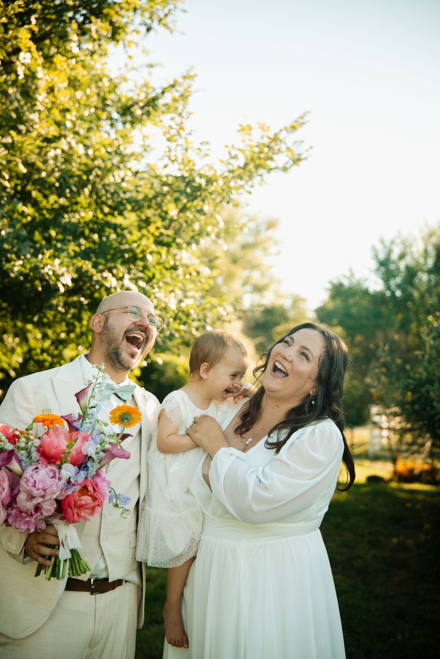 Laughing bride and groom in white wedding attire hold their baby at an outdoor summer wedding.