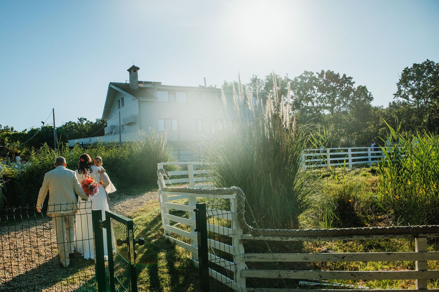 Couple walking toward a white barn venue surrounded by lush greenery and white fences on a sunny day.