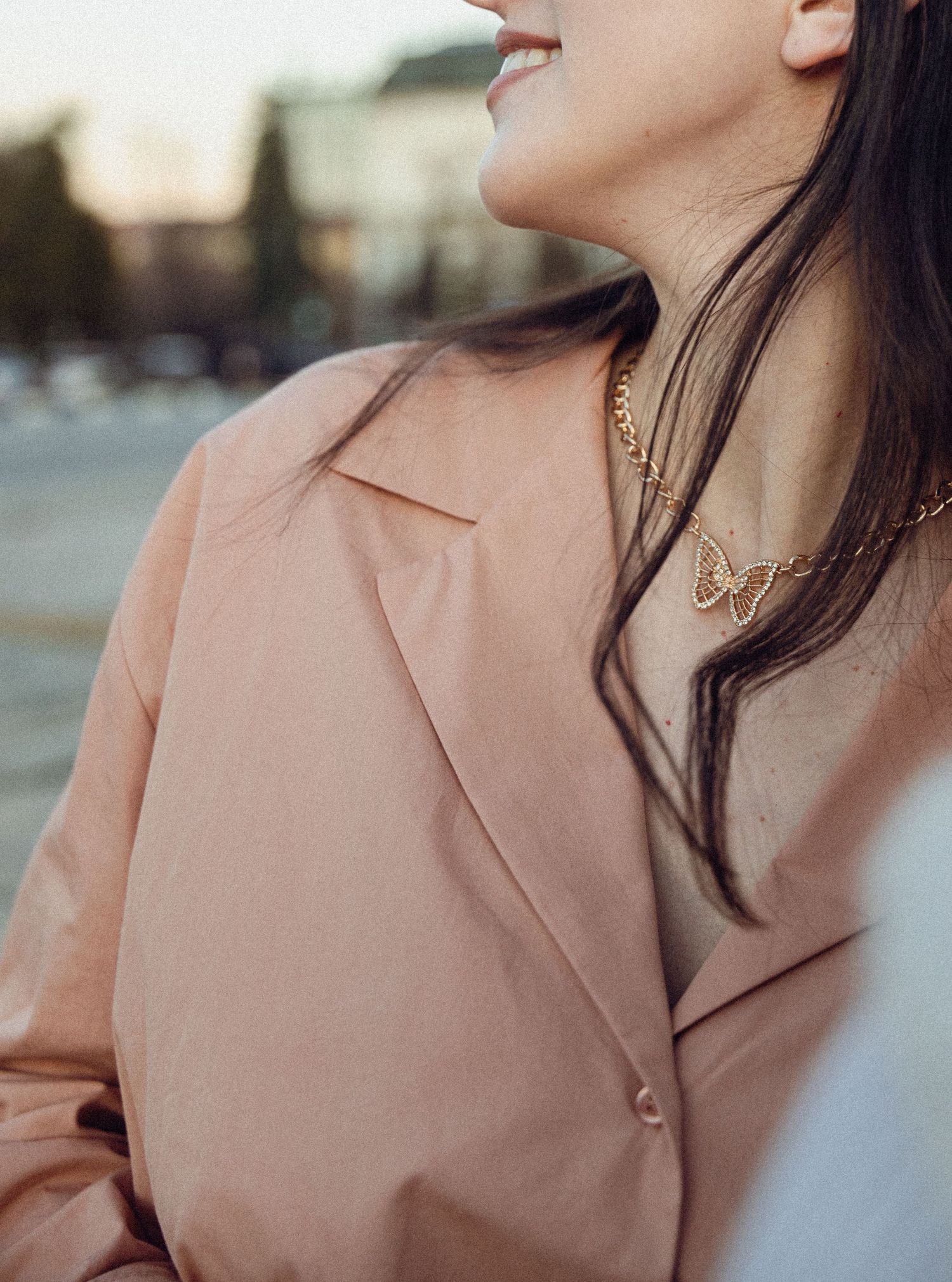 Close-up fashion detail of a beige blazer collar and gold chain necklace against a blurred urban background.