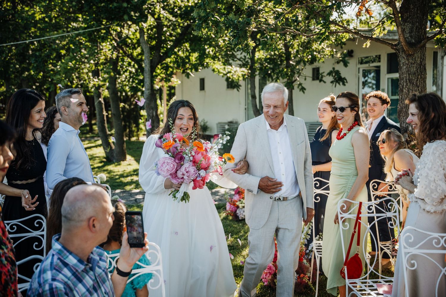 Bride in white gown holds colorful bouquet walking with guests at outdoor wedding ceremony surrounded by trees.