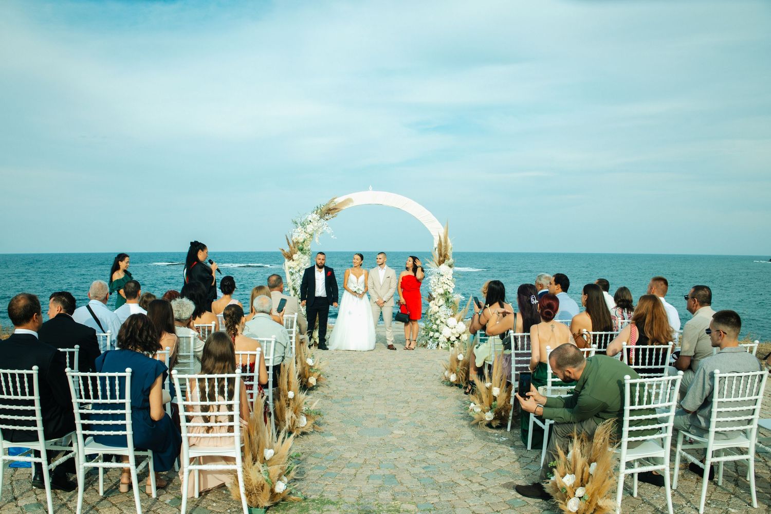 Beach wedding ceremony with couple at floral arch altar, guests seated in white chairs by the ocean.