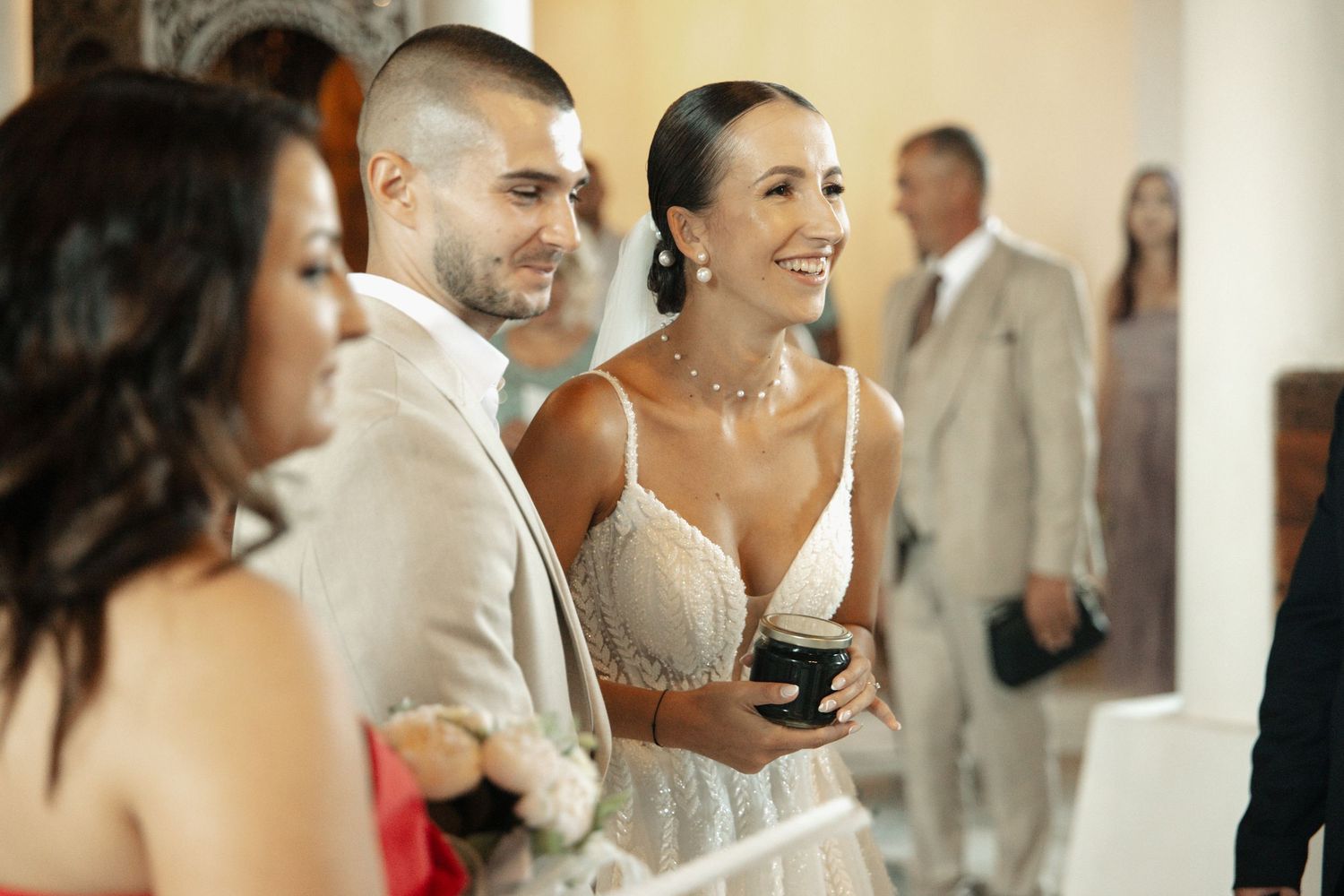 Smiling couple in wedding attire stand together during ceremony, bride in white gown, groom in light suit.