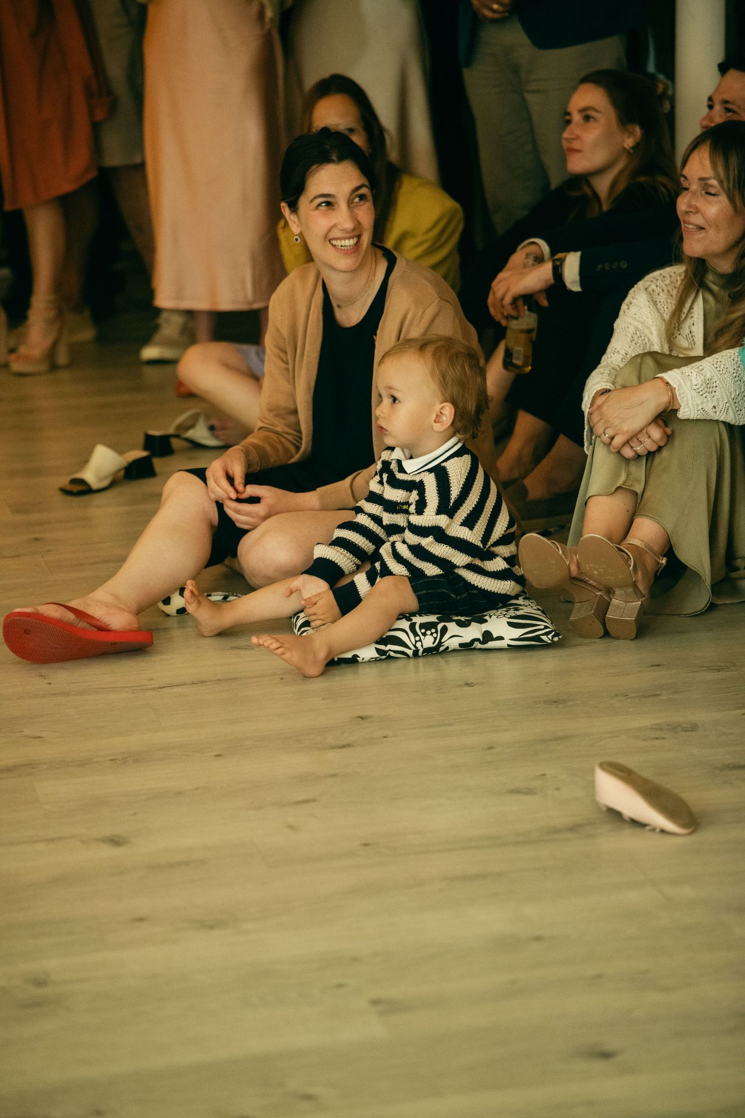 A smiling woman sits on a wooden floor with a baby in a striped outfit at a casual indoor gathering.