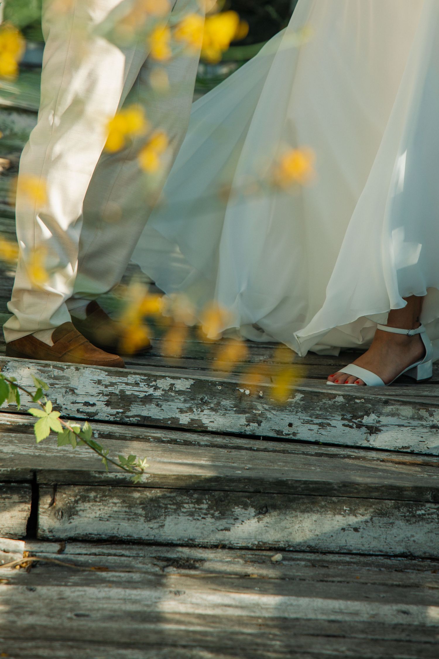 Close-up of bride and groom feet on rustic wooden dock, white wedding dress flowing among yellow wildflowers.