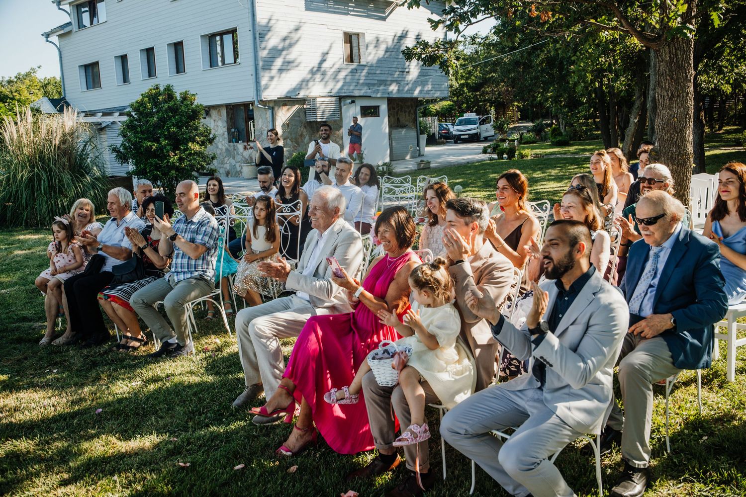 Wedding guests seated on chairs applaud during an outdoor garden ceremony beside a white estate building.