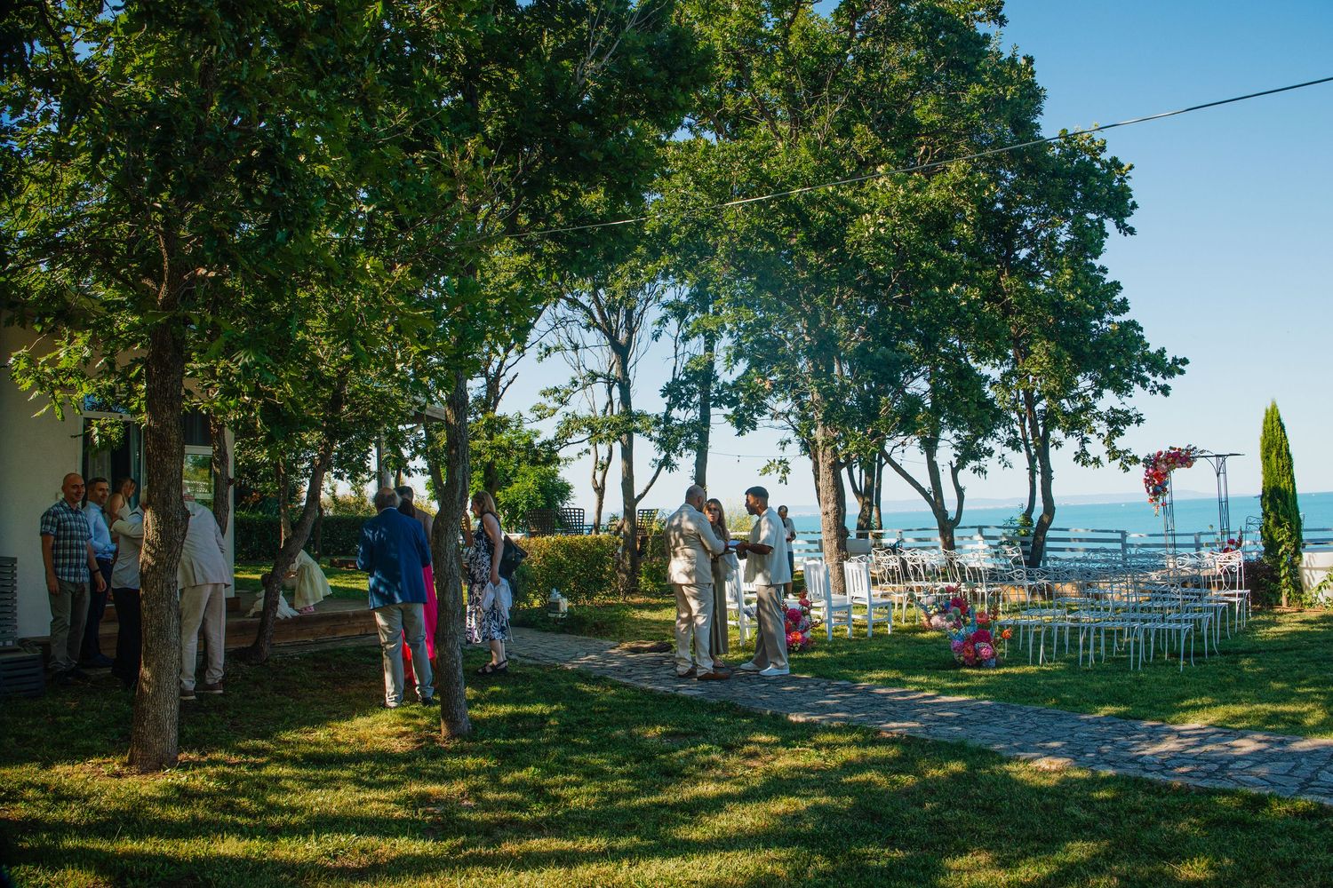 Outdoor wedding ceremony on lush green lawn with guests gathered among tall trees overlooking a scenic waterfront.