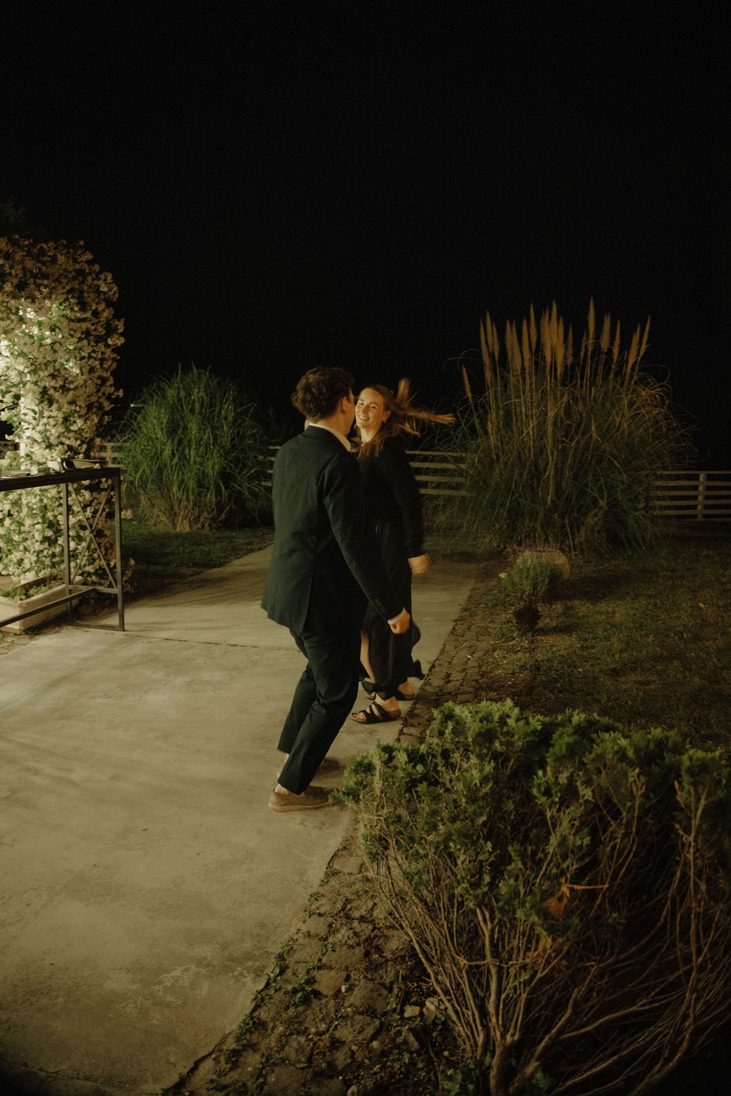 Couple dancing outdoors at night in garden with ornamental grasses and pathway lighting.