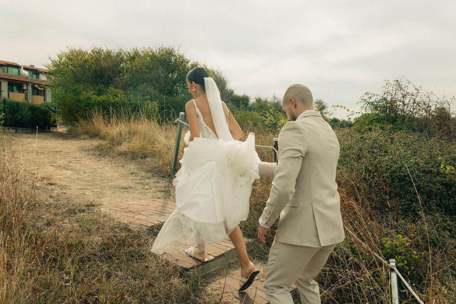 Bride and groom walking hand in hand along a scenic dirt path surrounded by wild greenery on their wedding day.