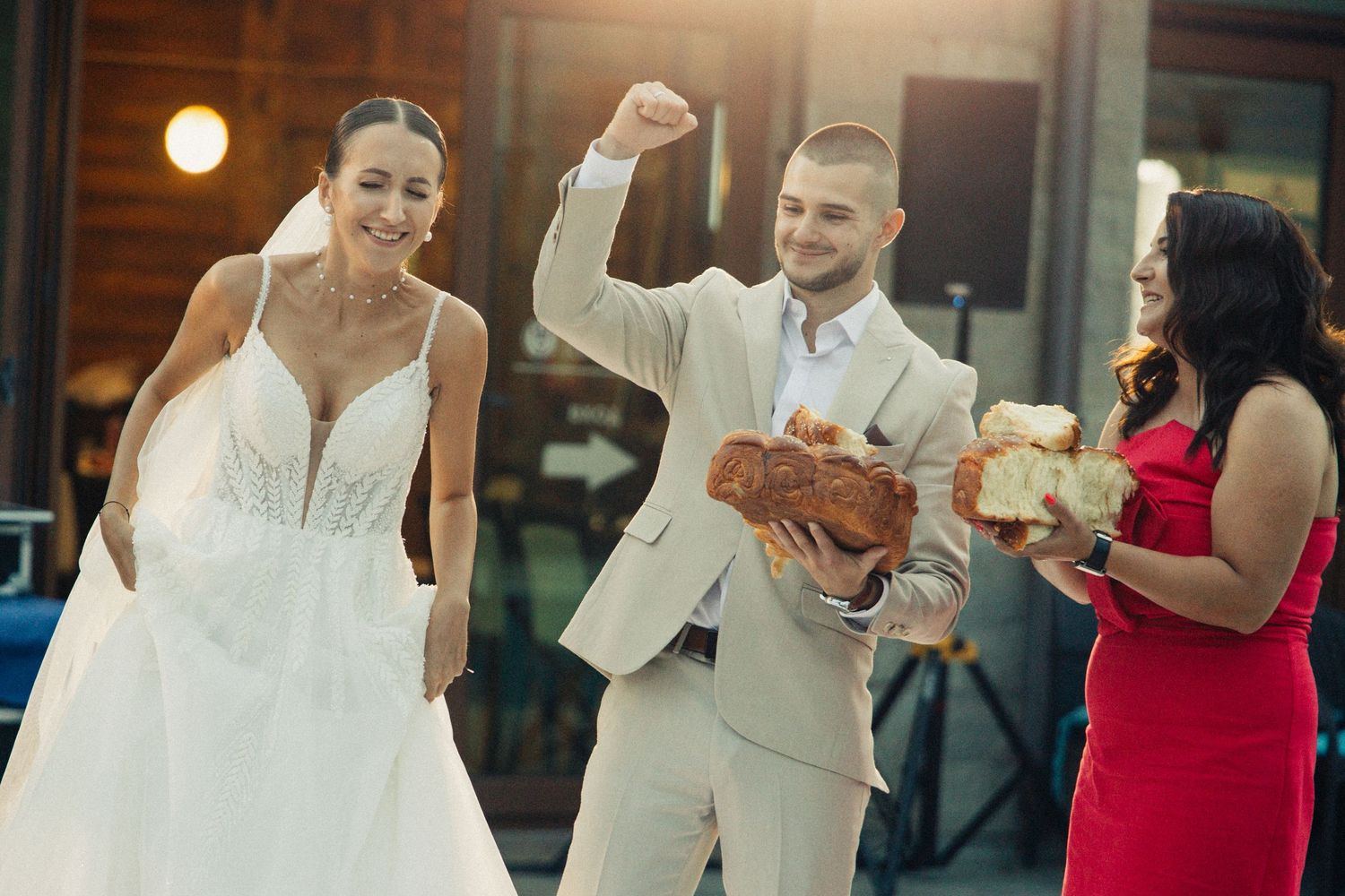 Groom in beige suit celebrates holding bread at wedding reception while bride and guest in red dress look on.