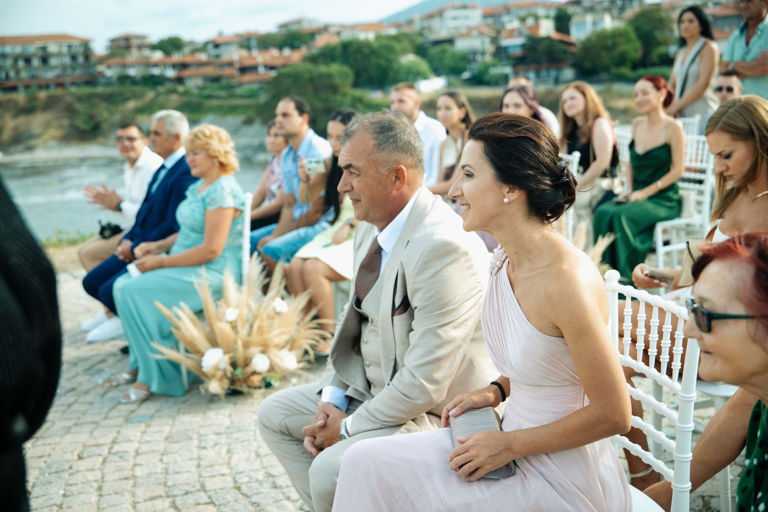 Smiling bride in white dress seated with groom in tan suit at an outdoor waterfront wedding ceremony.