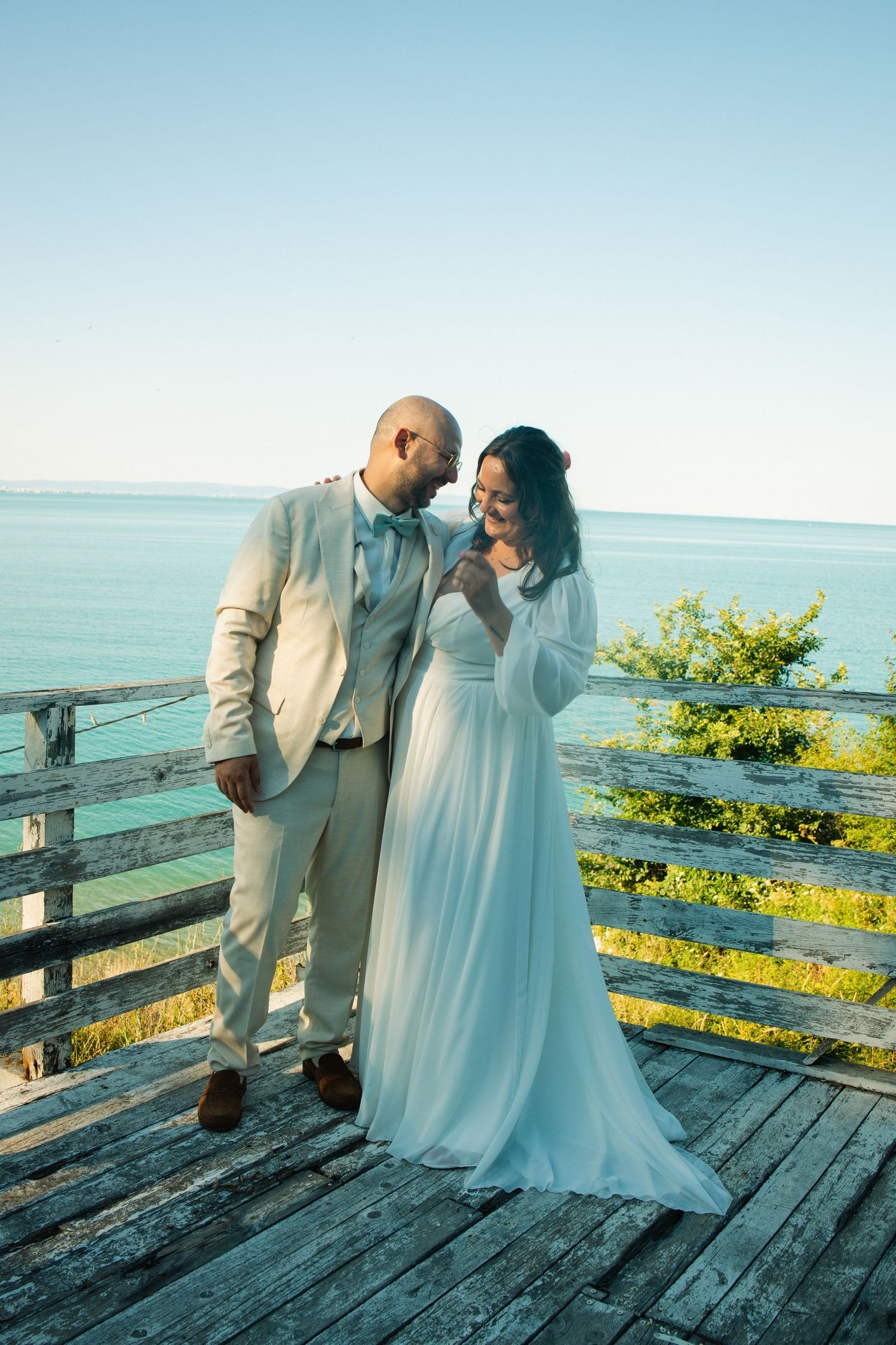 Bride and groom share a romantic moment on a wooden deck overlooking turquoise ocean waters on their wedding day.