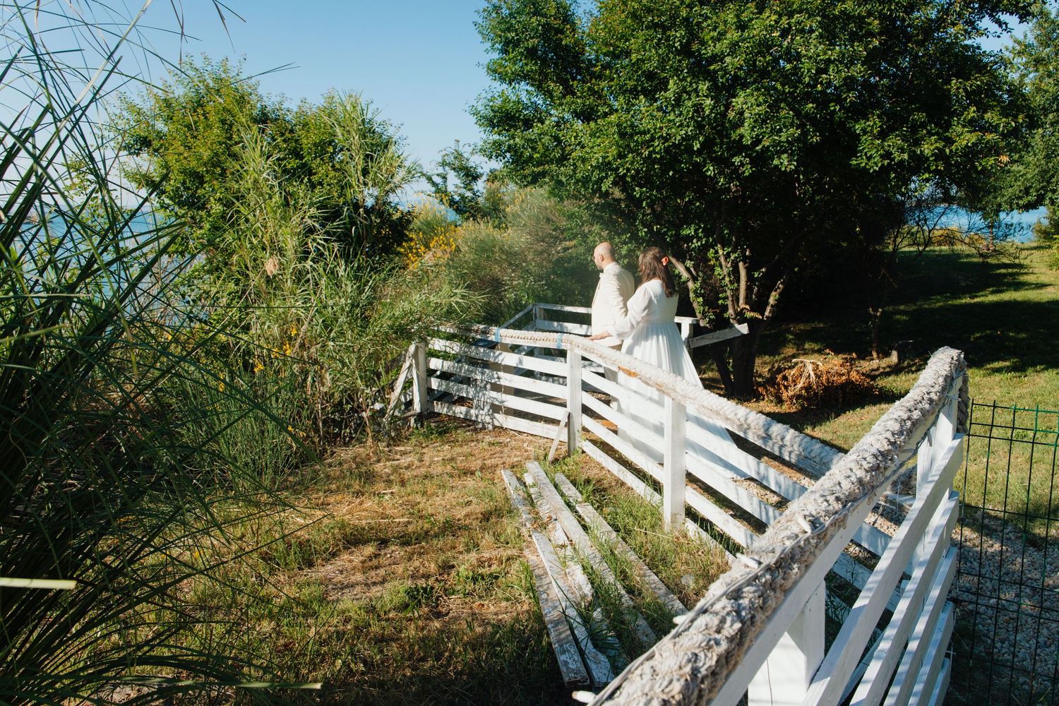 Bride and groom share a romantic moment on a white wooden fence bridge surrounded by lush green trees.