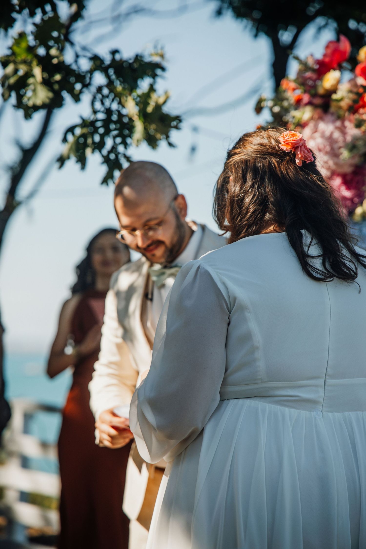 Groom smiling while exchanging vows with bride at an outdoor wedding ceremony by the sea.