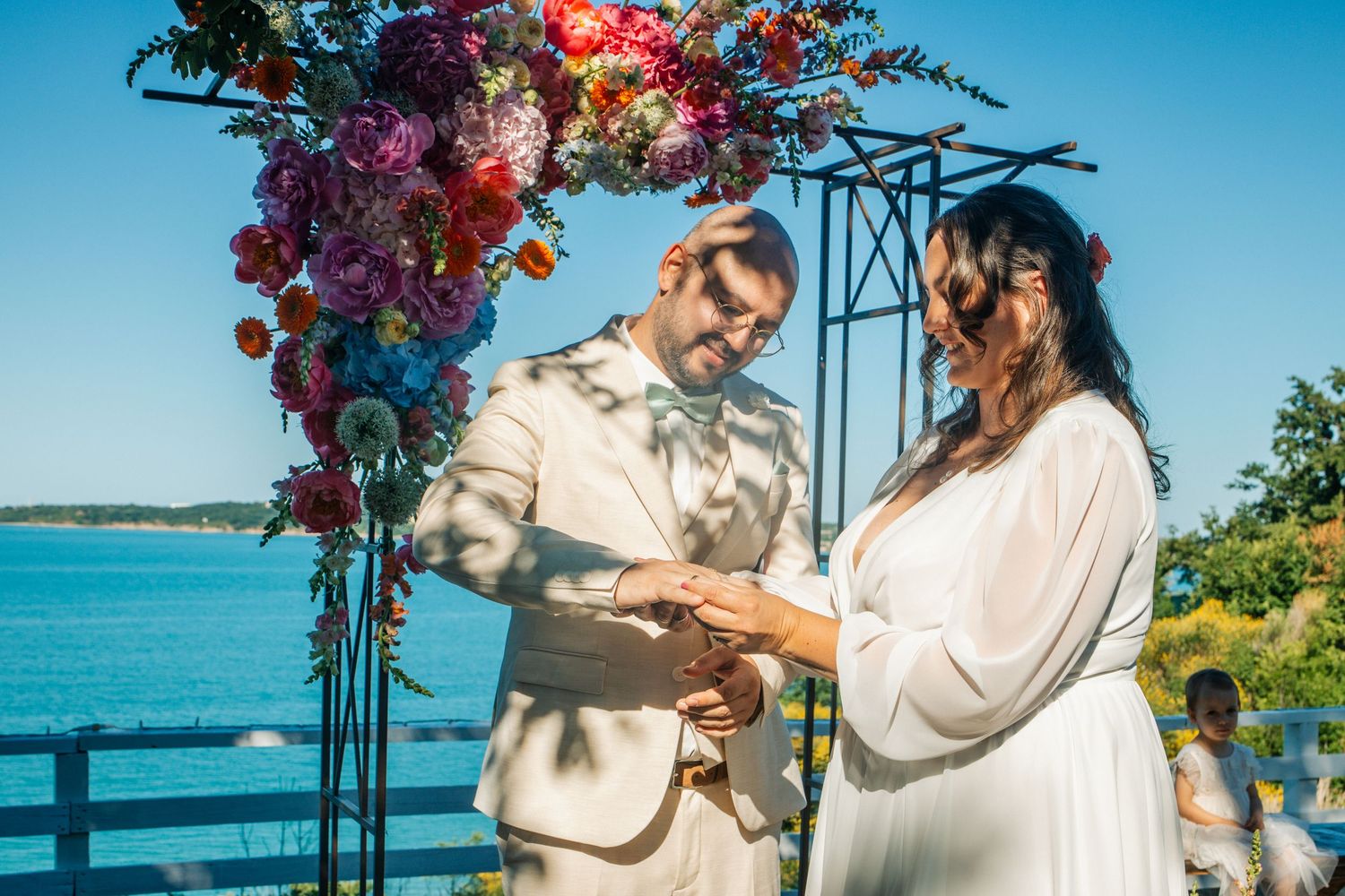 Couple exchanging rings at outdoor wedding ceremony under colorful floral arch with blue water in background.