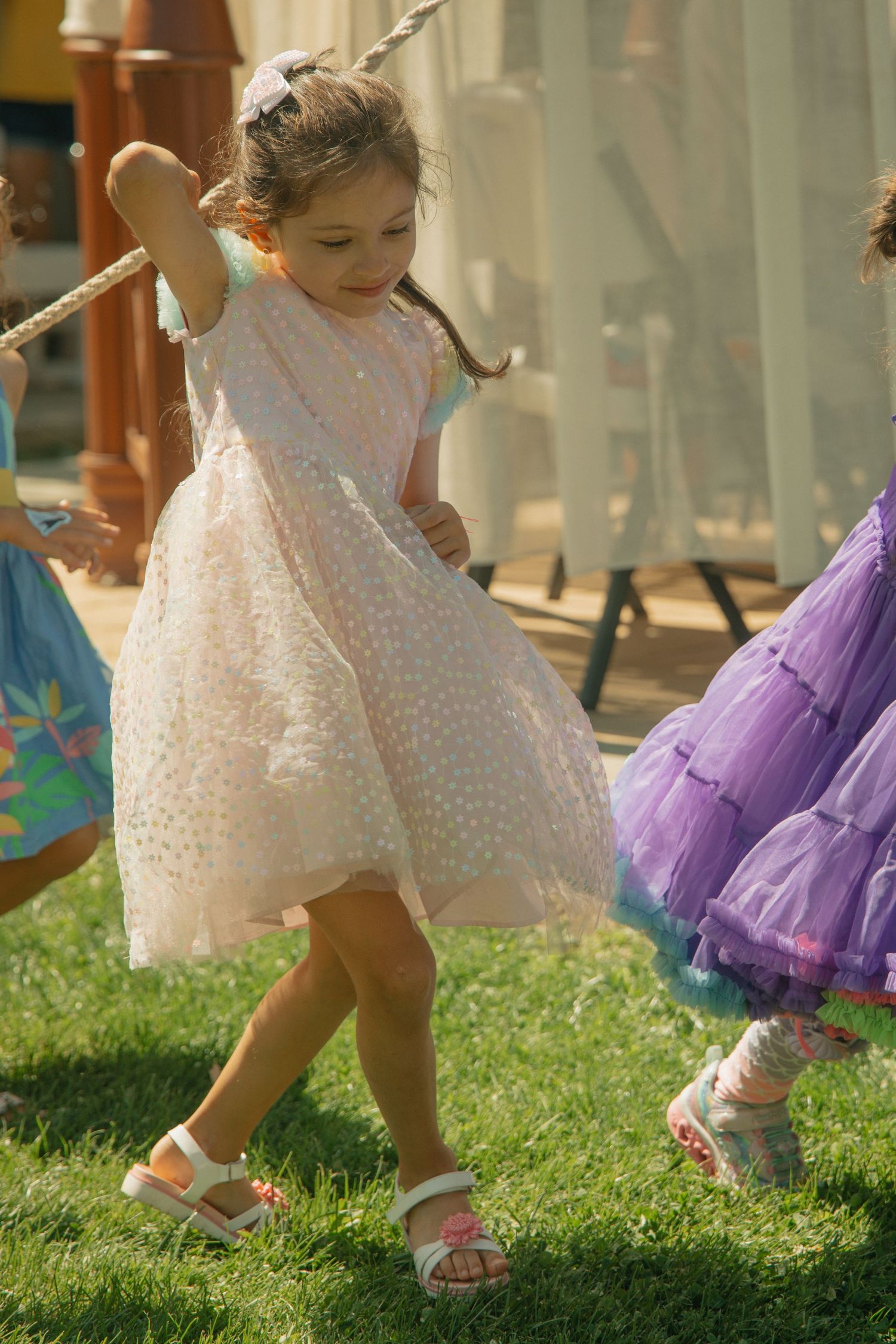 Young girl in white lace dress and sandals dancing on green grass at outdoor party.