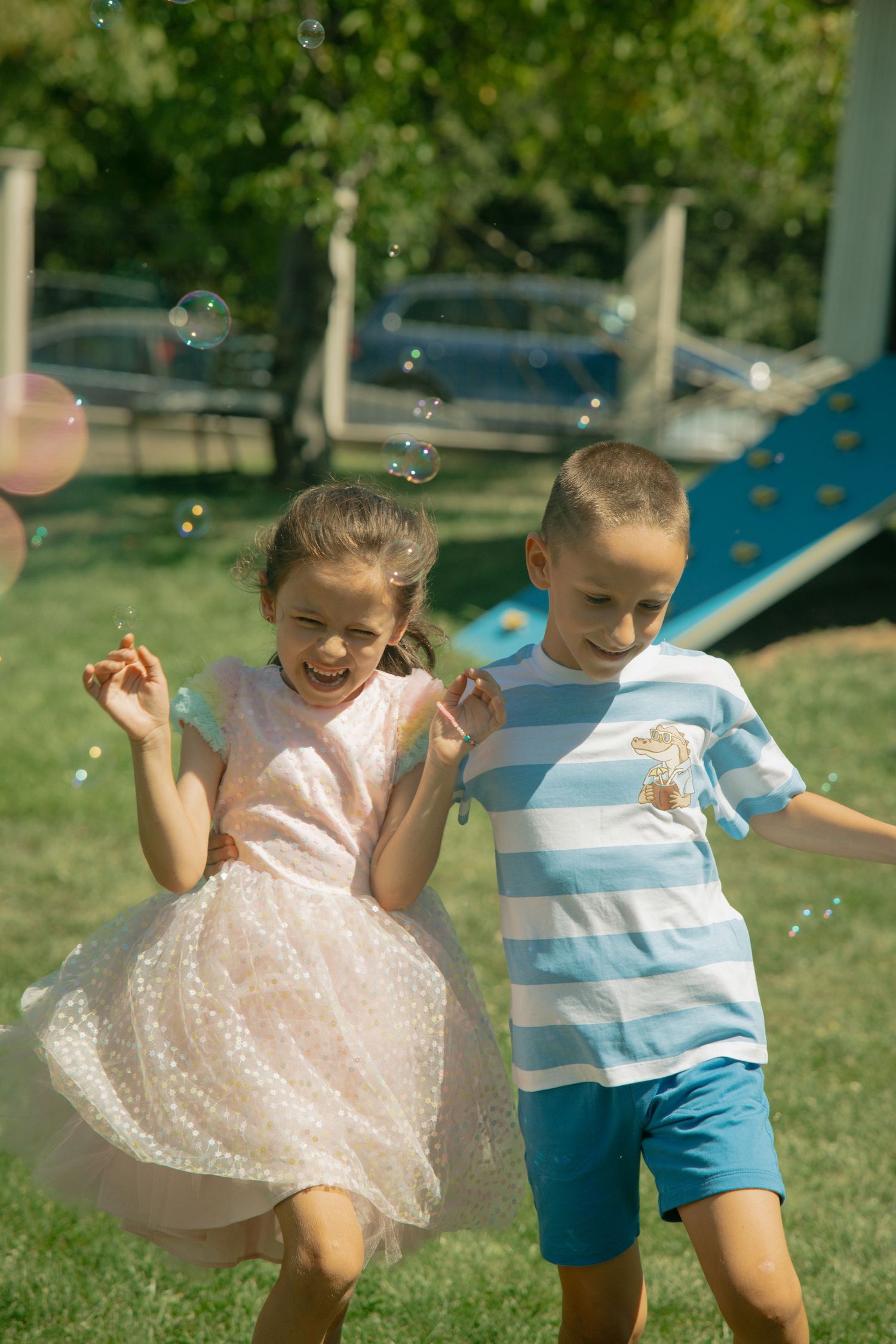 Two happy children playing with soap bubbles outdoors in a sunny park, girl in pink dress and boy in striped shirt.