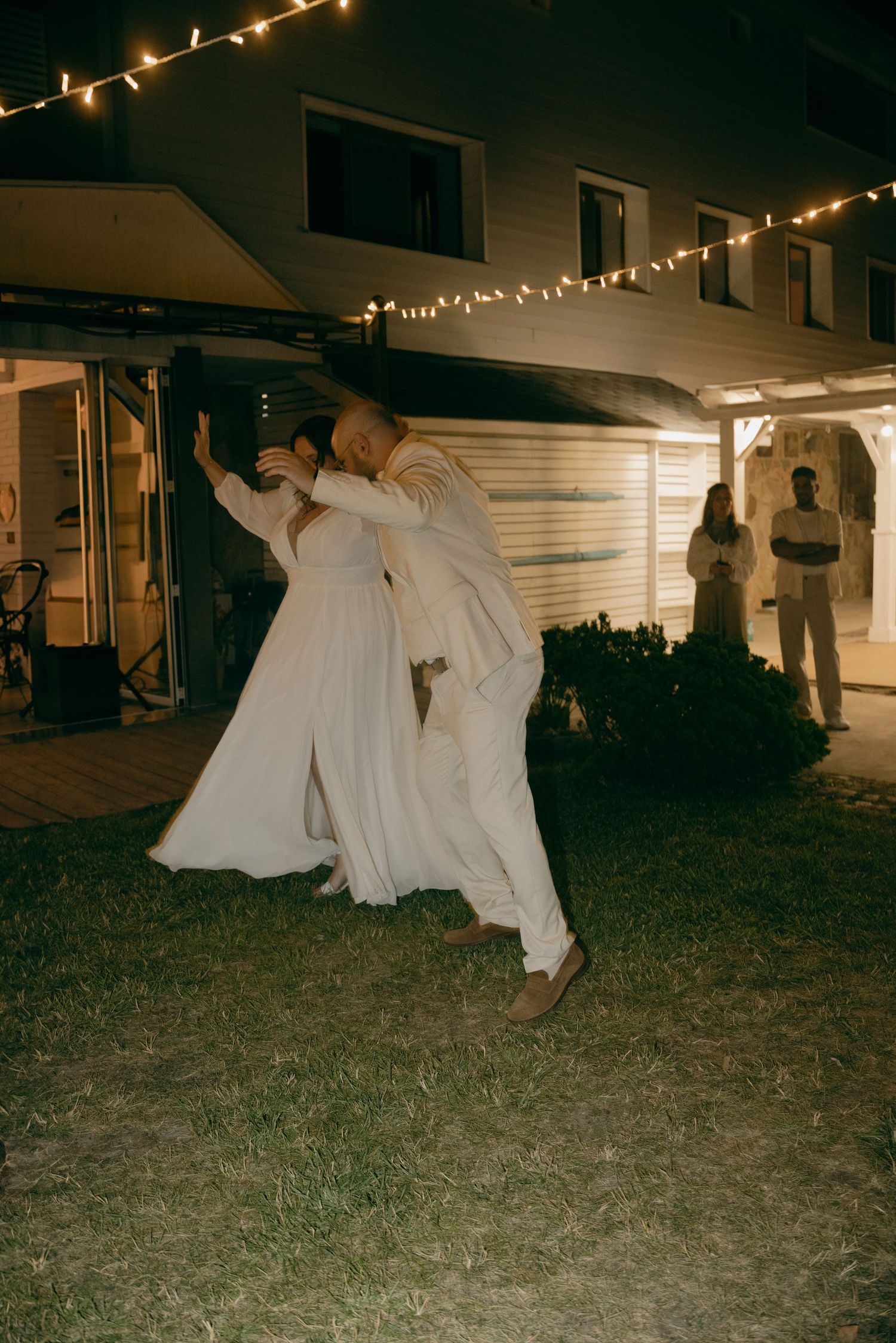 Bride and groom share a romantic dip dance outdoors at night under string lights during their wedding reception.
