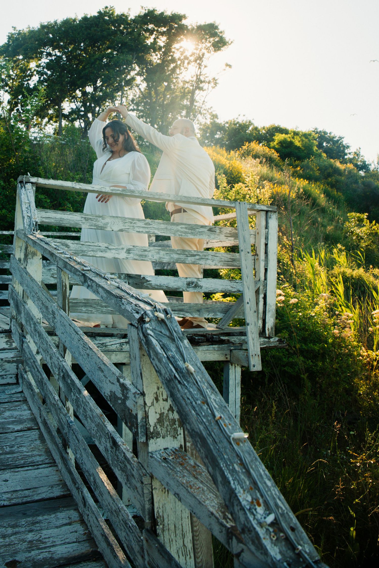 Bride and groom standing on rustic wooden bridge at sunset, bathed in golden light with lush green foliage.
