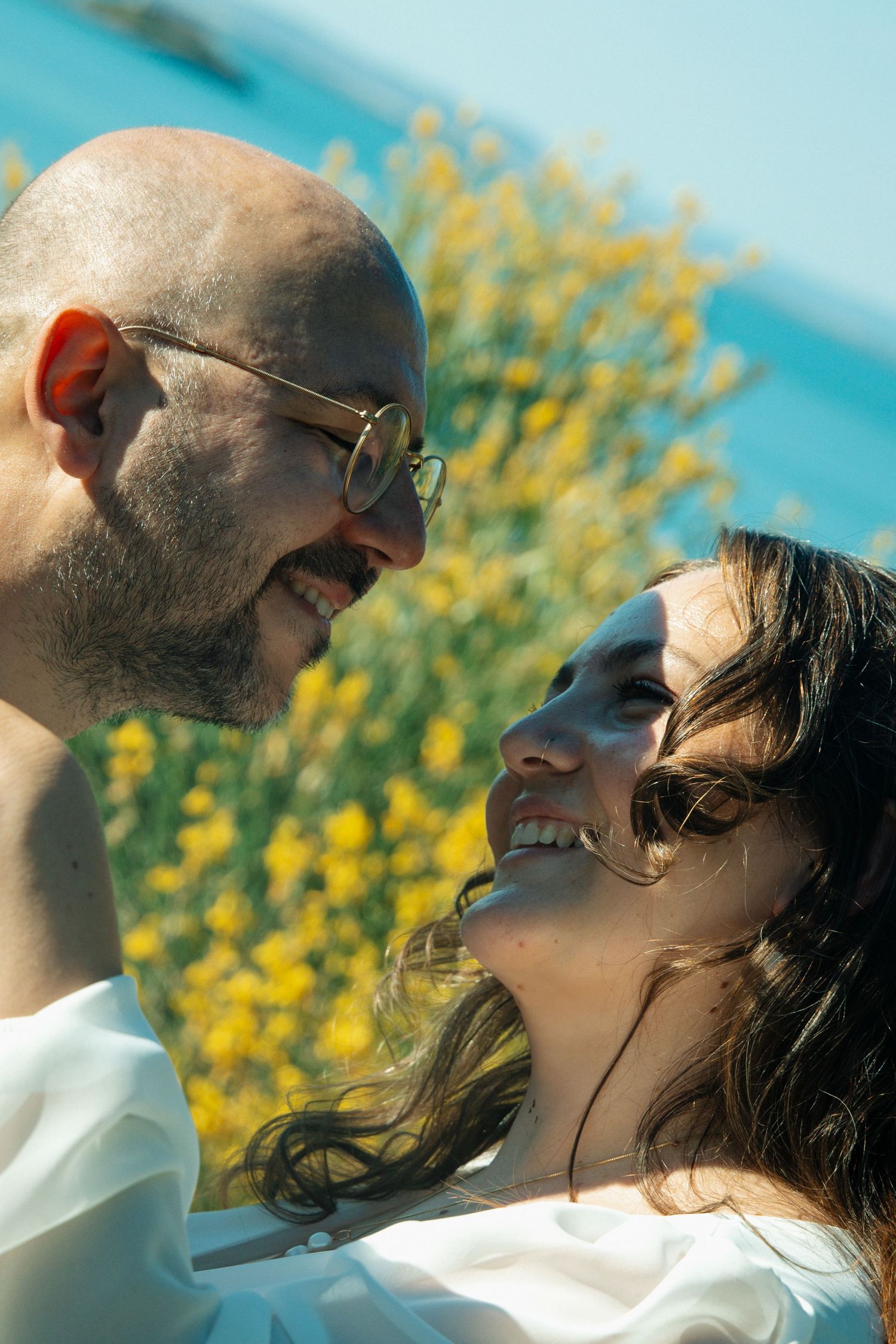 Happy couple smiling at each other outdoors with yellow flowers and blue water in the background on a sunny day.