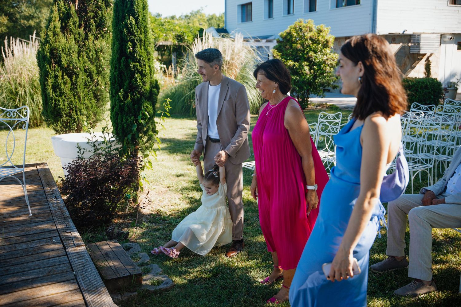 Guests in elegant attire walk through a garden wedding venue with cypress trees and white chairs.