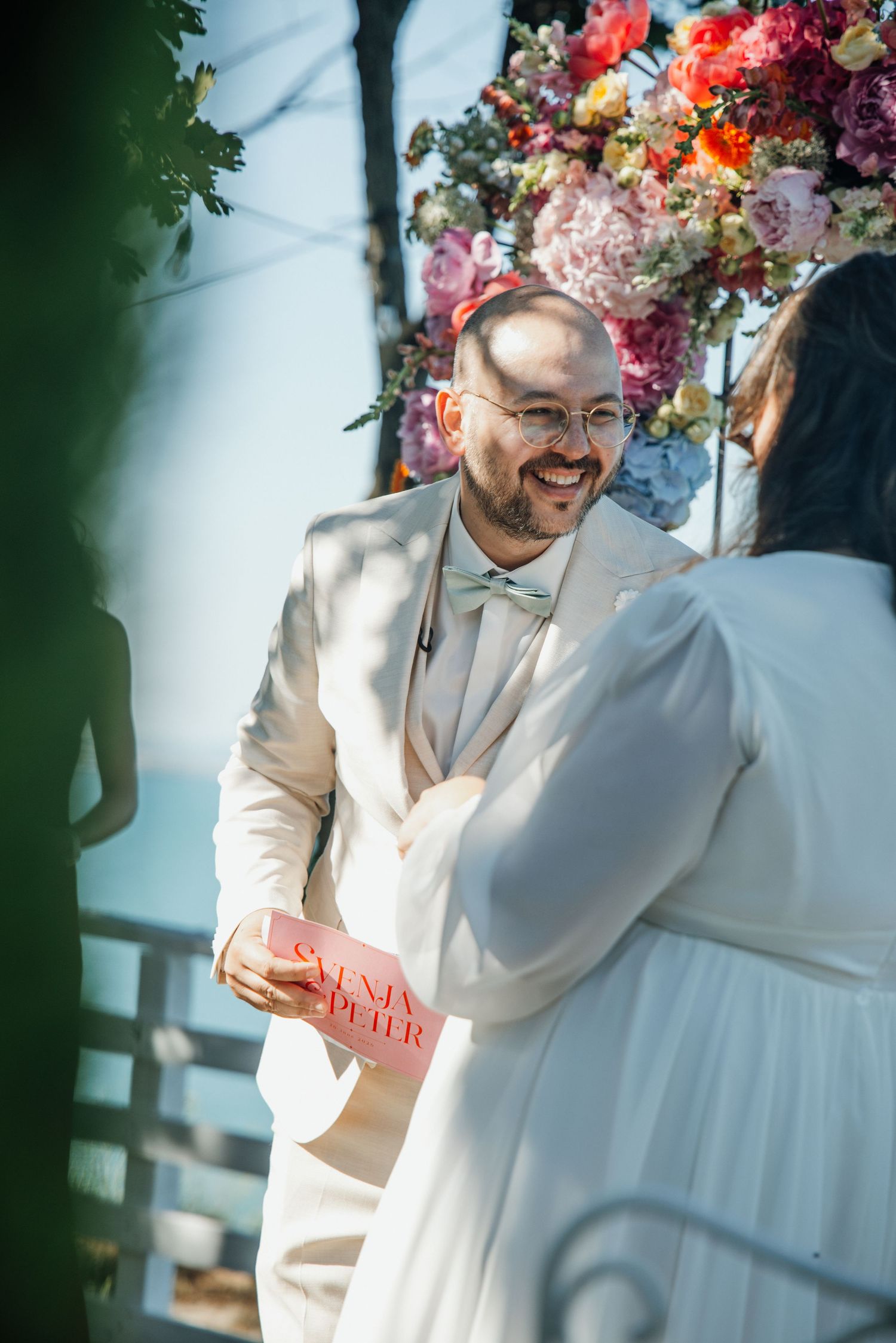 Smiling groom in white suit holding vow book at colorful floral altar during outdoor ceremony.