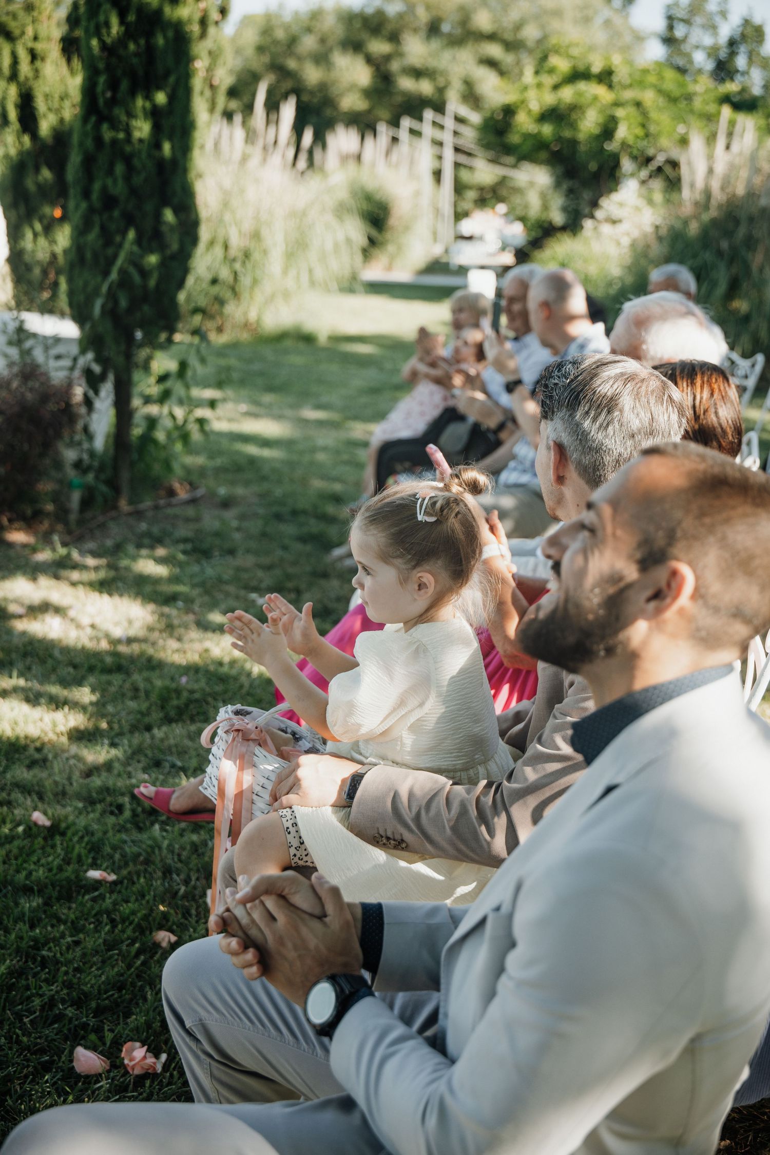 Wedding guests seated outdoors with a young child clapping during a sunny garden ceremony.
